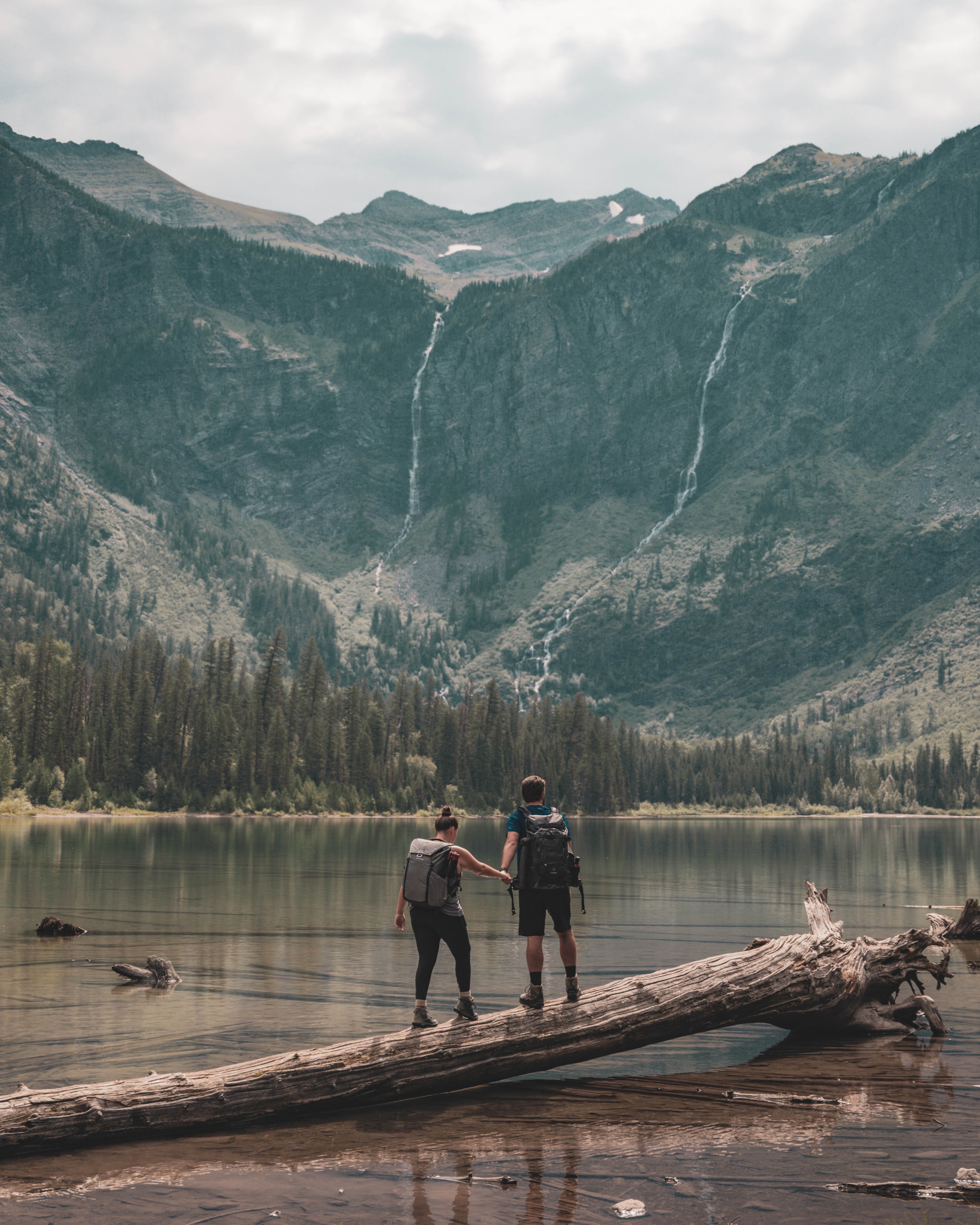 Two people stand on a log by a lake with mountains and waterfalls in the background.