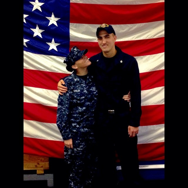 Two people in military uniforms standing in front of an American flag, one person looking at the other.