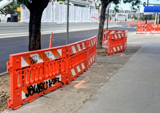Orange construction barriers along sidewalk with trees and street in background, some graffiti on barriers, daytime.
