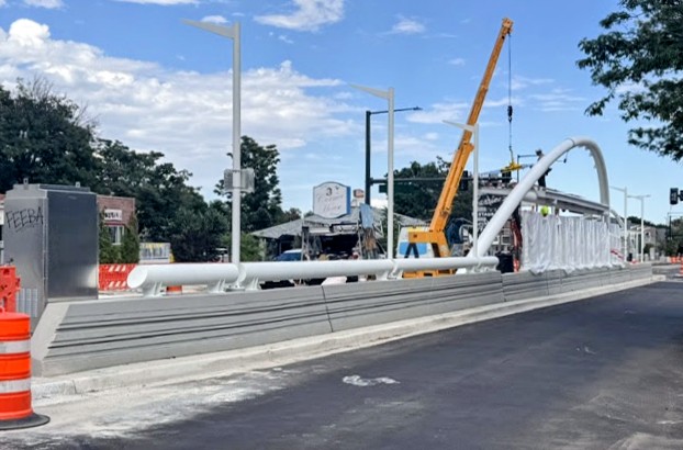 Construction site with crane, pipes, and barriers on a street, blue sky with clouds, trees in background.