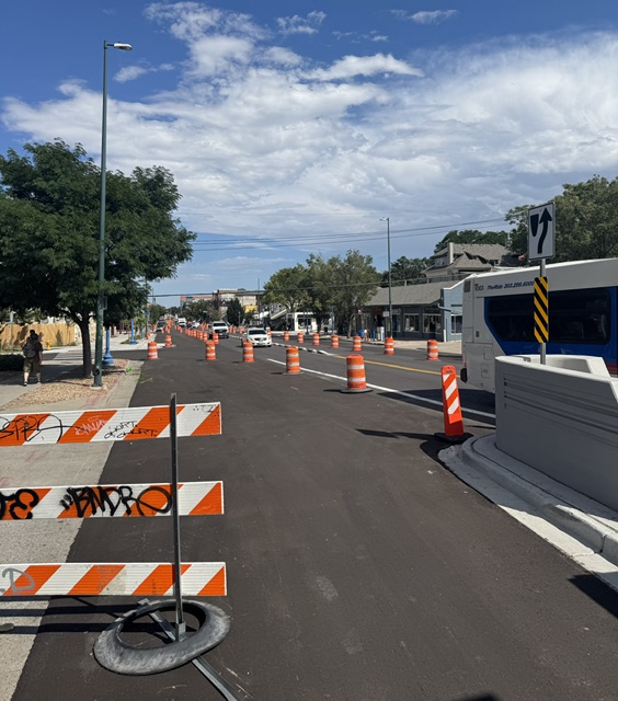 Street under construction with orange traffic cones and barriers, blue sky with clouds, trees, and buildings in background.