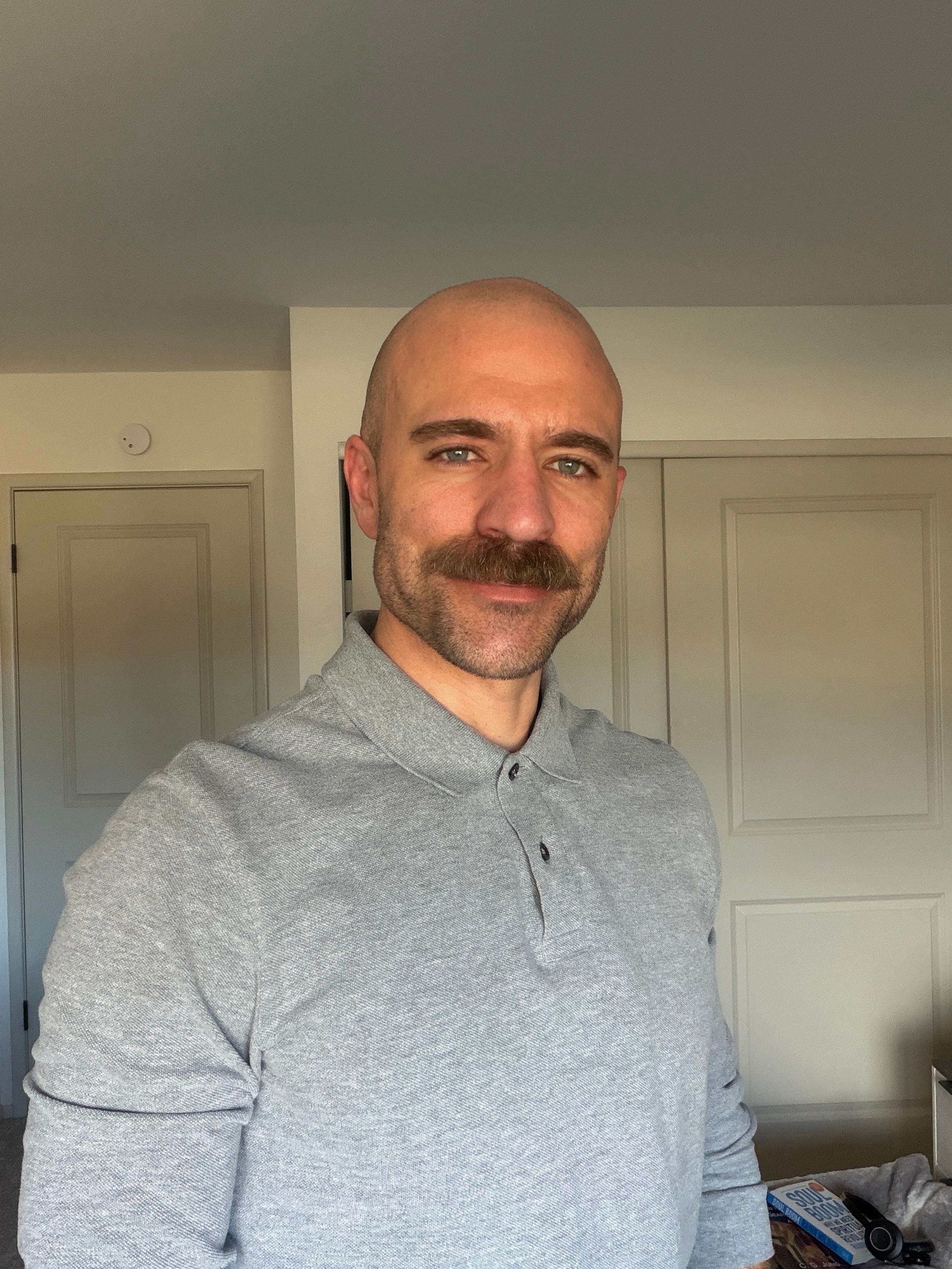 Bald man with a mustache wearing a gray polo shirt, smiling indoors with closed doors in the background.