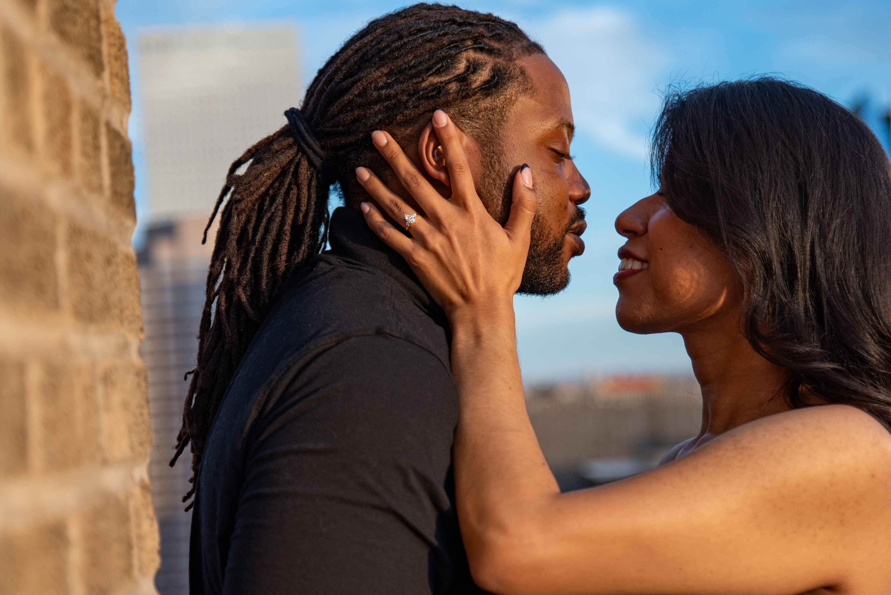 A man and woman face each other closely, with the woman's hand on the man's face, outdoors with a cityscape background.
