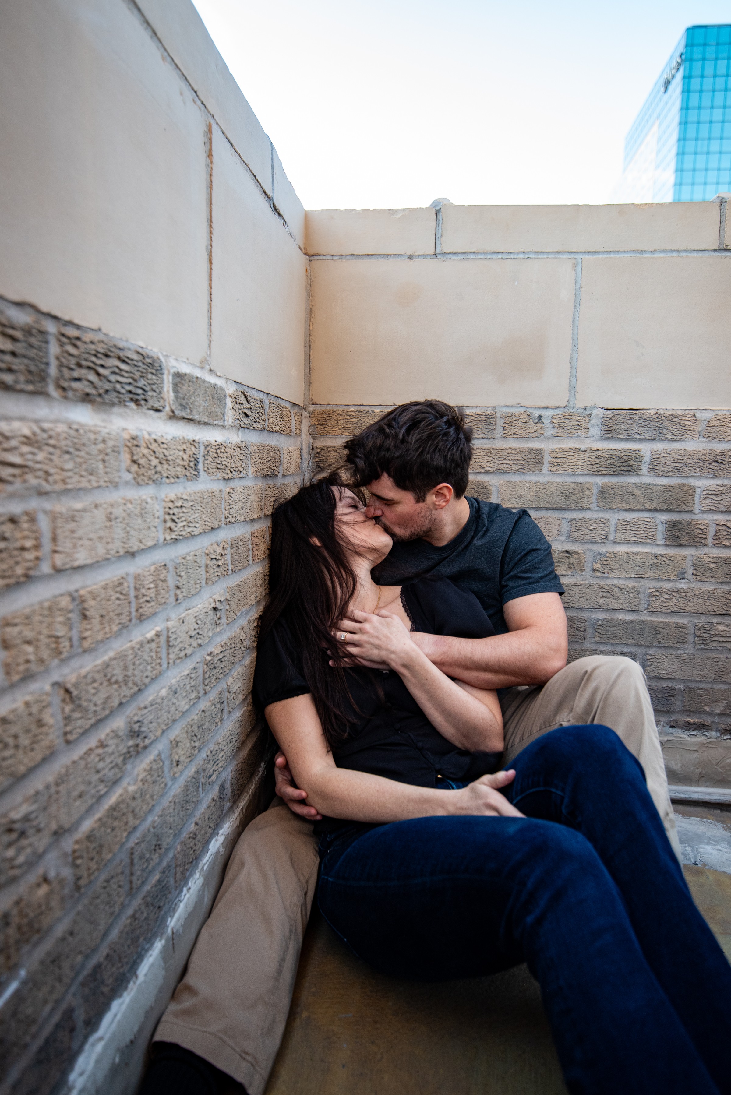 Two people sitting closely in a corner, kissing, with brick and stone walls behind them.