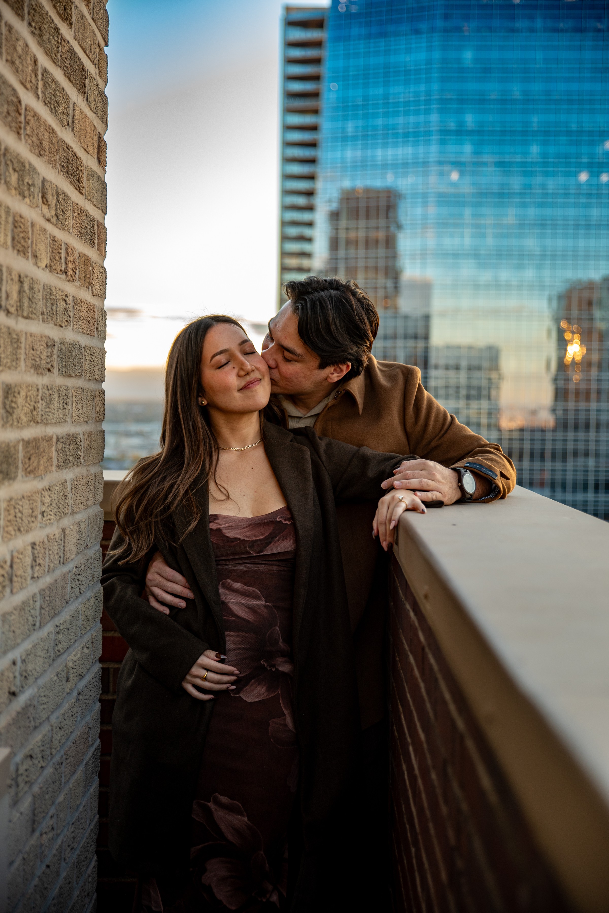 A couple stands on a balcony with city buildings in the background, embracing and smiling at each other.