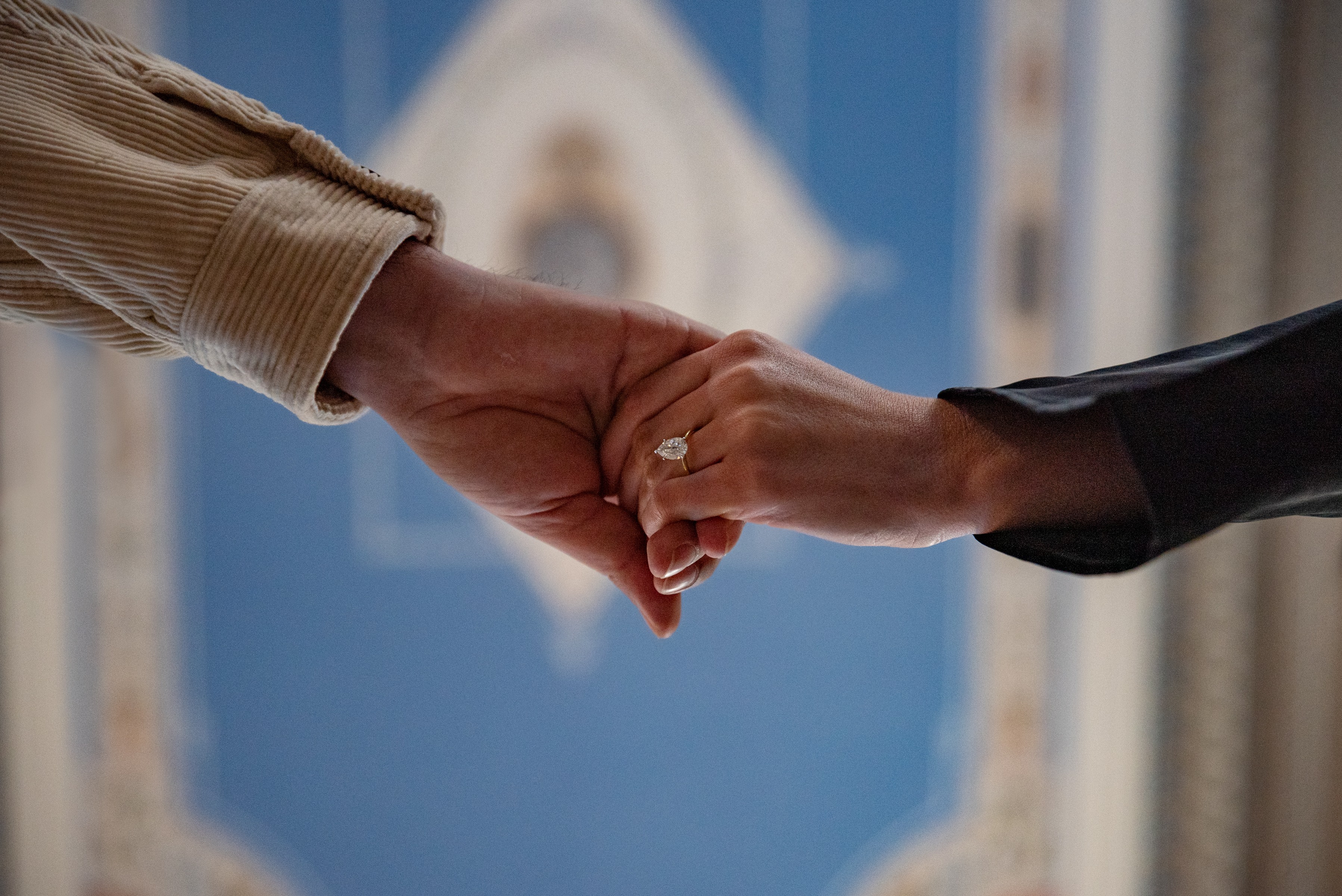 Two people hold hands, one with a beige sleeve and the other with a black sleeve, against a blurred blue background.