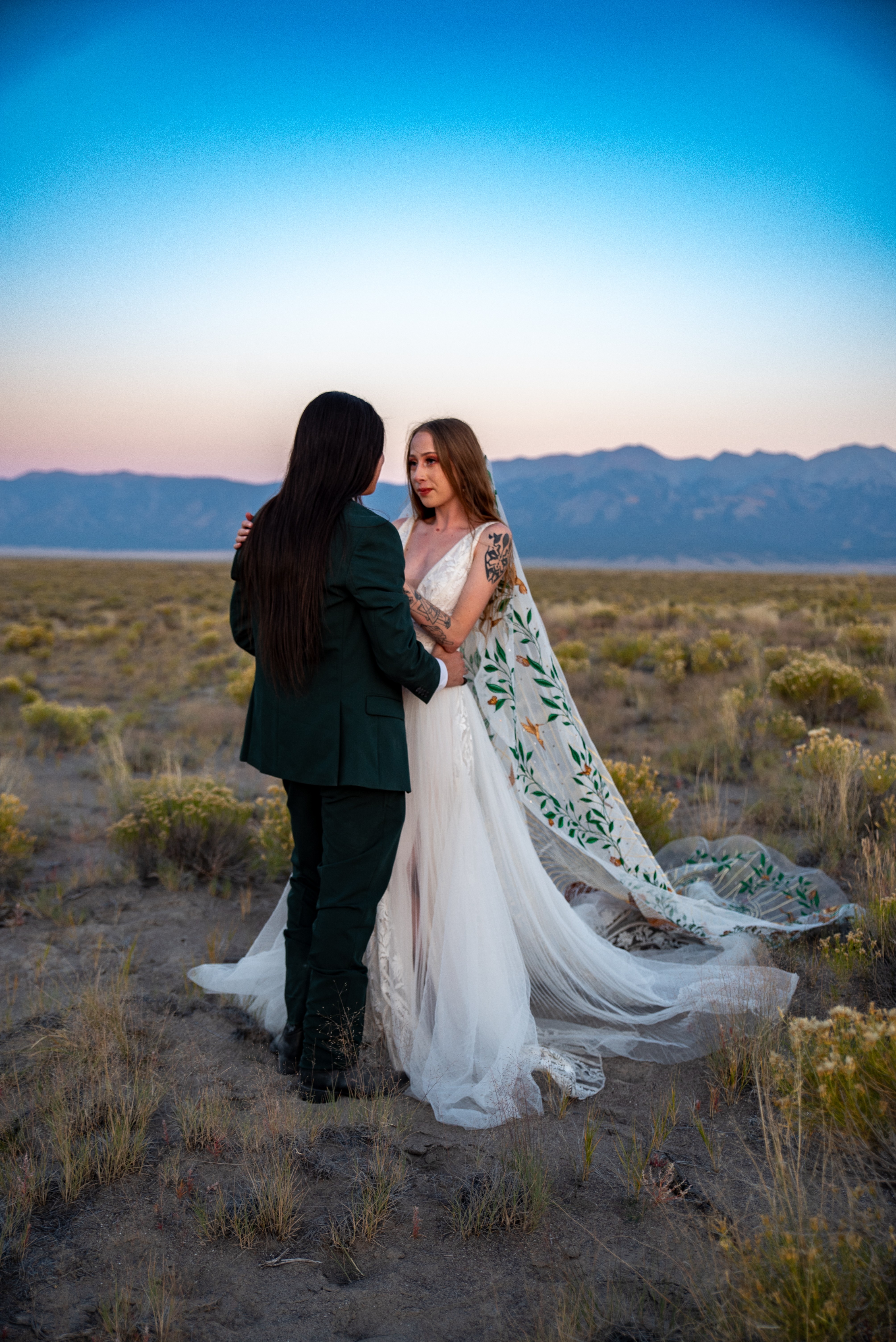 Two women in wedding attire standing in a desert landscape with mountains and a colorful sky in the background.