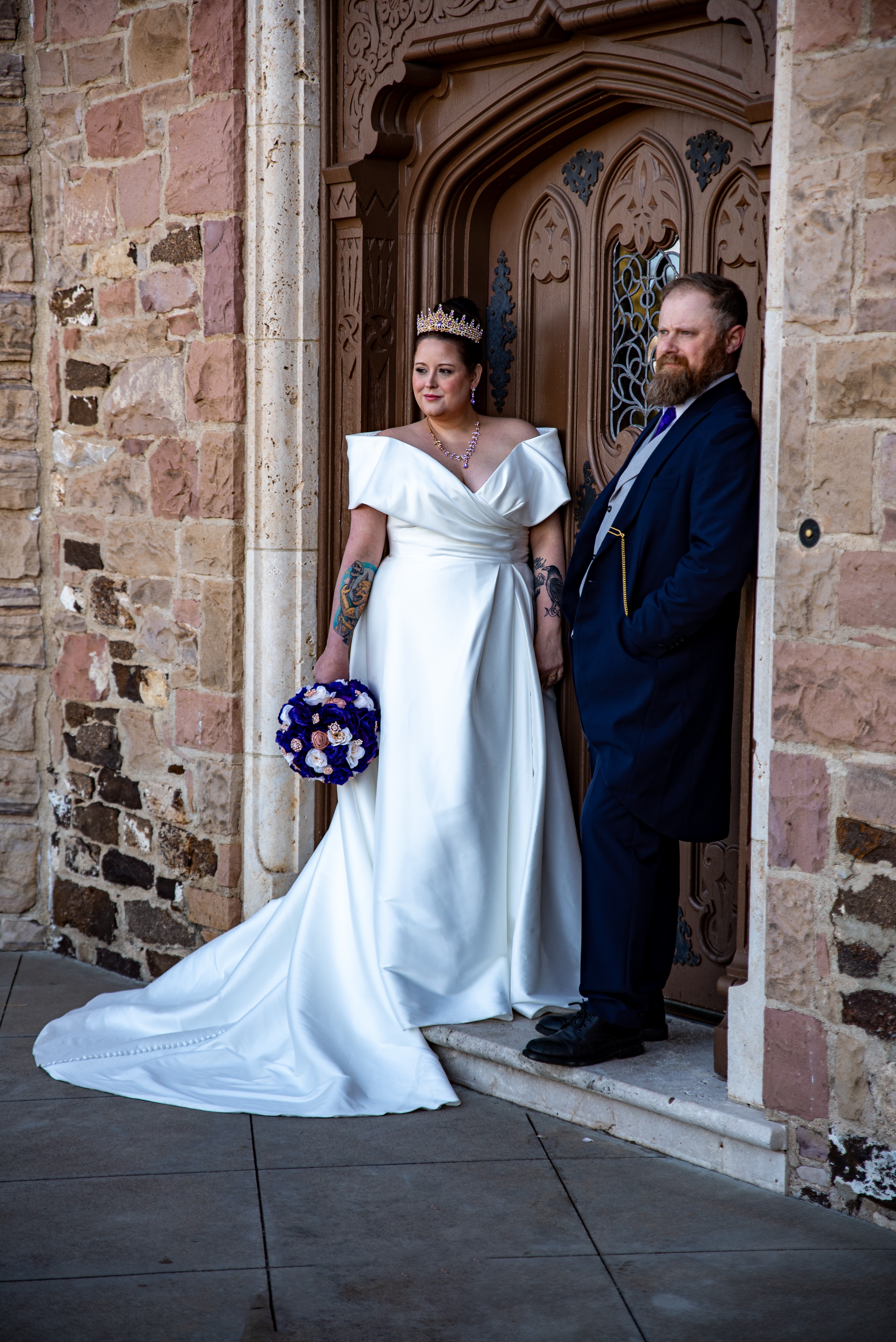 Bride and groom standing in front of a wooden door, bride holding a bouquet, both dressed in wedding attire.