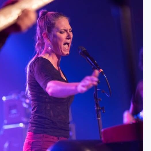 Woman singing passionately into a microphone on stage with blue lighting, wearing a black shirt and red pants.