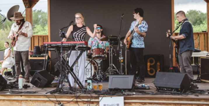 Five musicians perform on an outdoor stage with instruments and equipment, surrounded by trees and a cloudy sky.