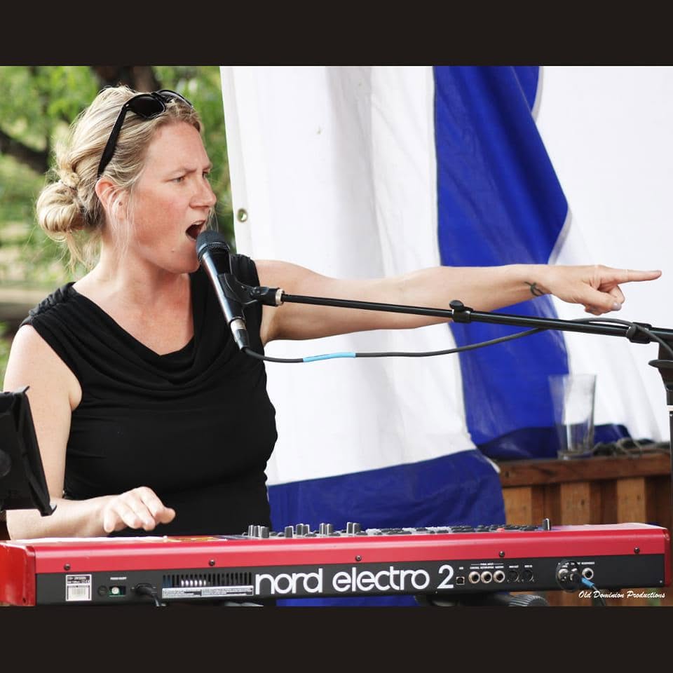 Woman singing into microphone while playing a red Nord Electro keyboard outdoors, with a blue and white backdrop.