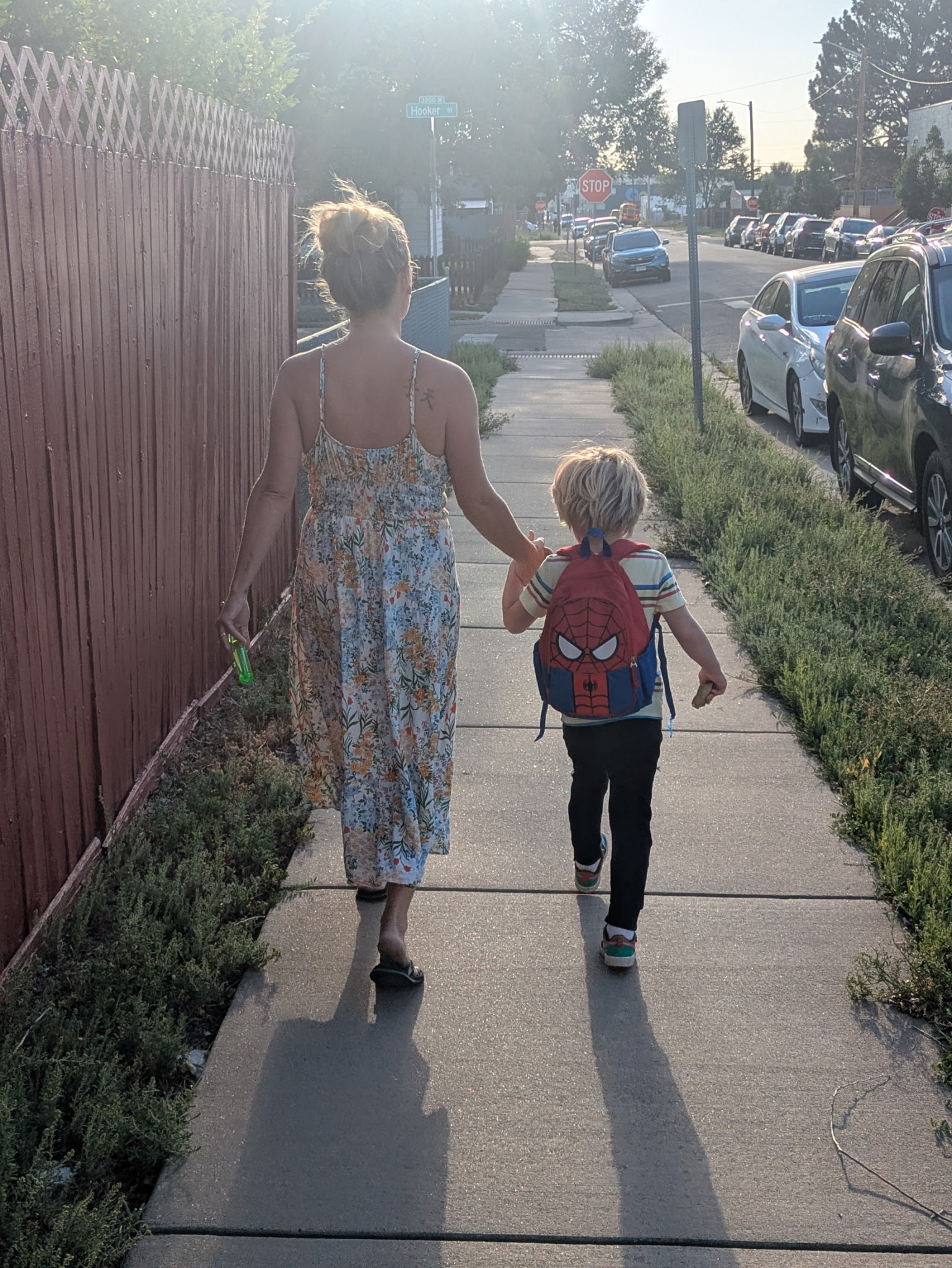 Woman and child walking hand in hand on sidewalk, with parked cars and trees in background, sunlight shining through.