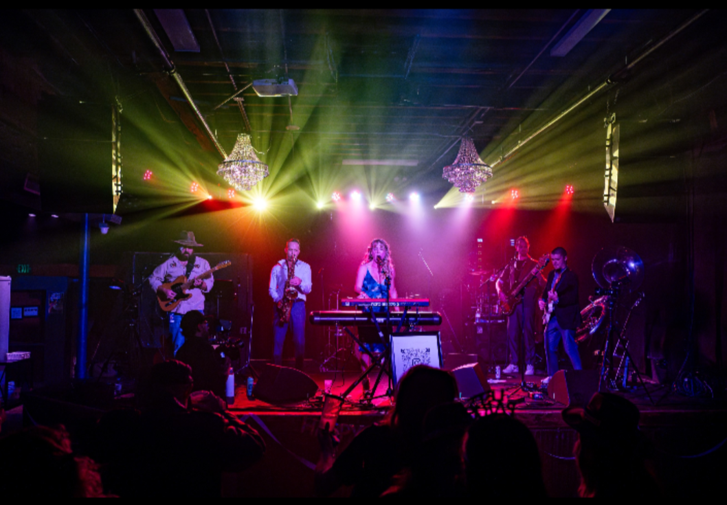 Band performs on stage with colorful lighting and chandeliers, audience visible in foreground.