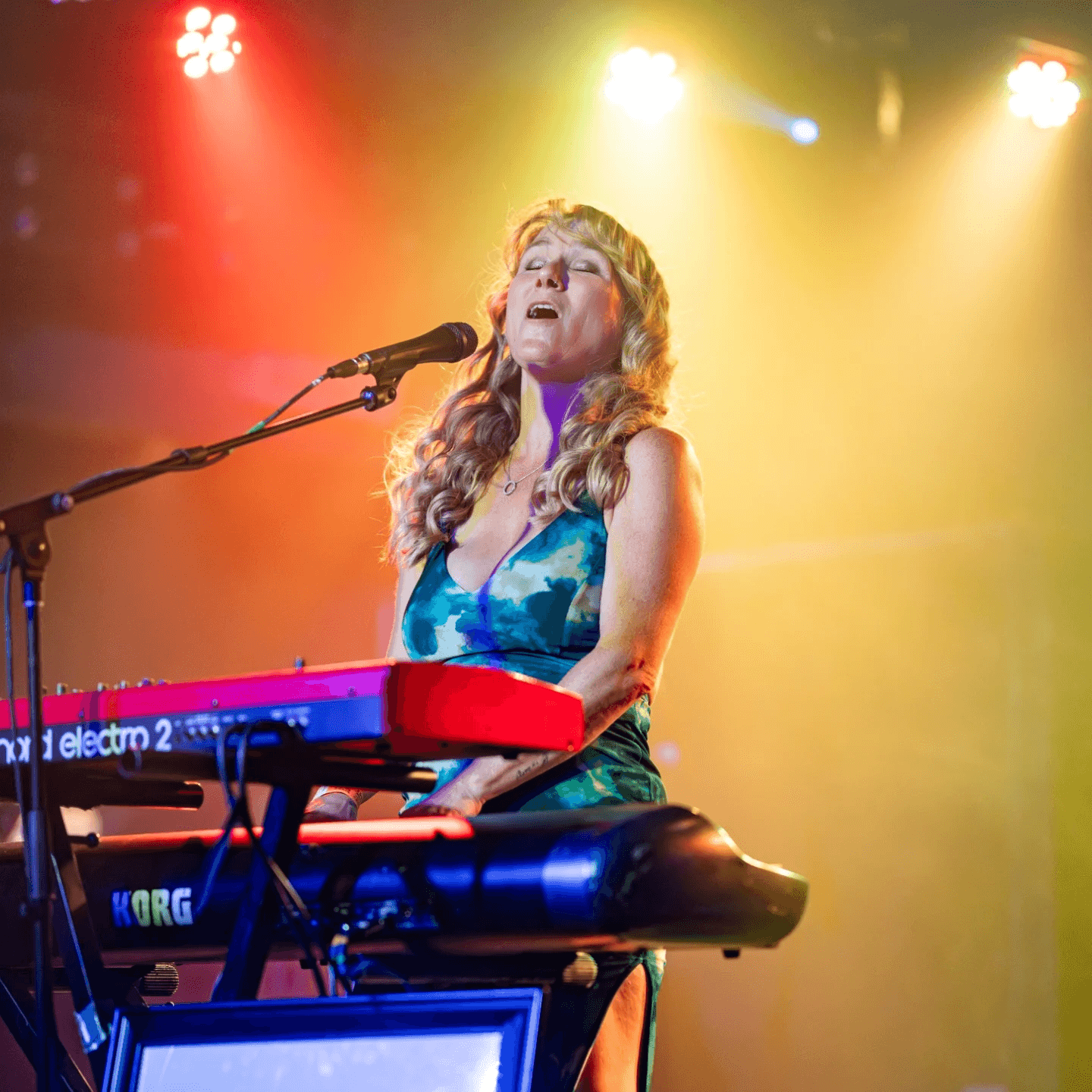Woman singing and playing a red keyboard on stage with colorful lights behind her.