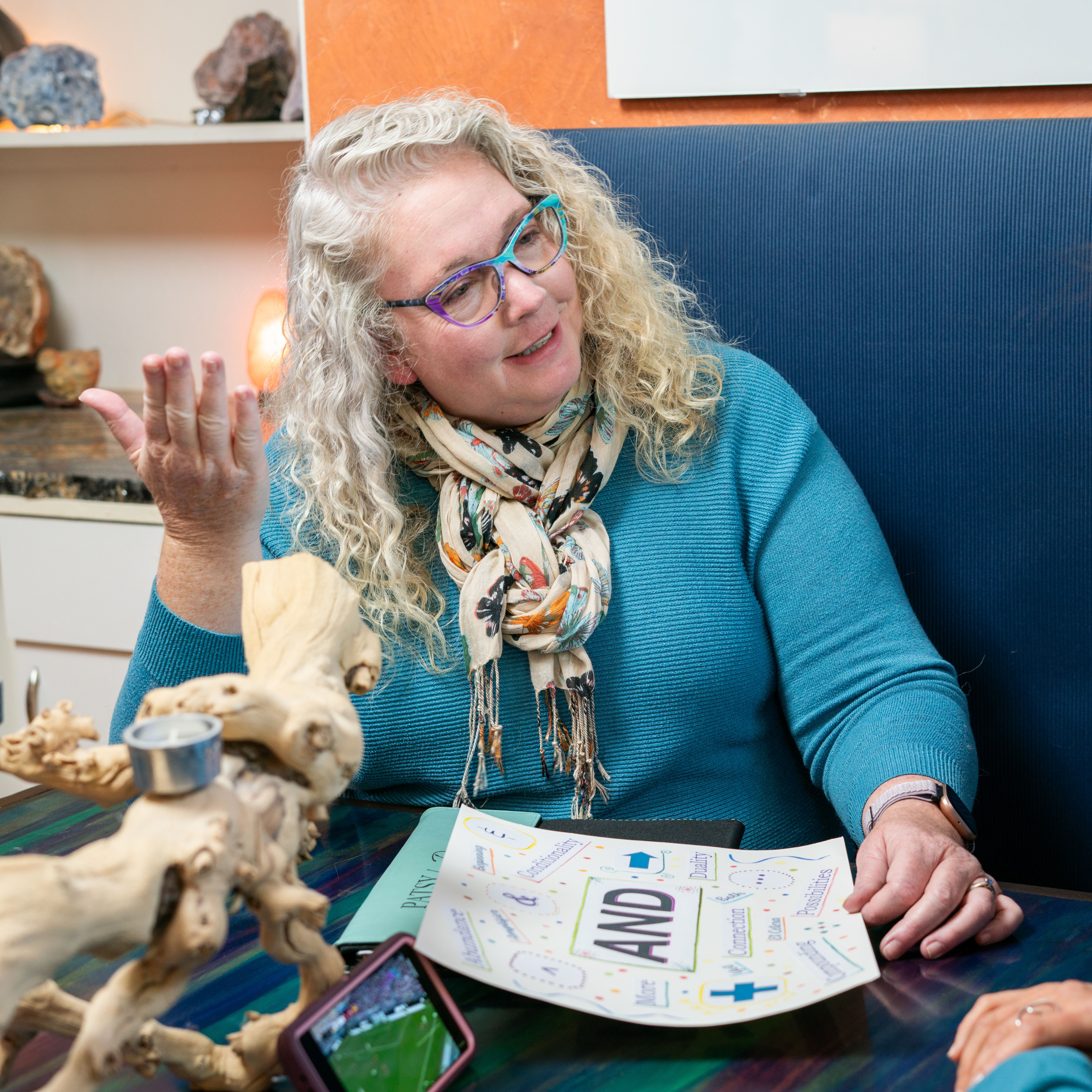 Woman with curly gray hair and glasses, wearing a blue sweater and patterned scarf, gesturing with her hand at a table.