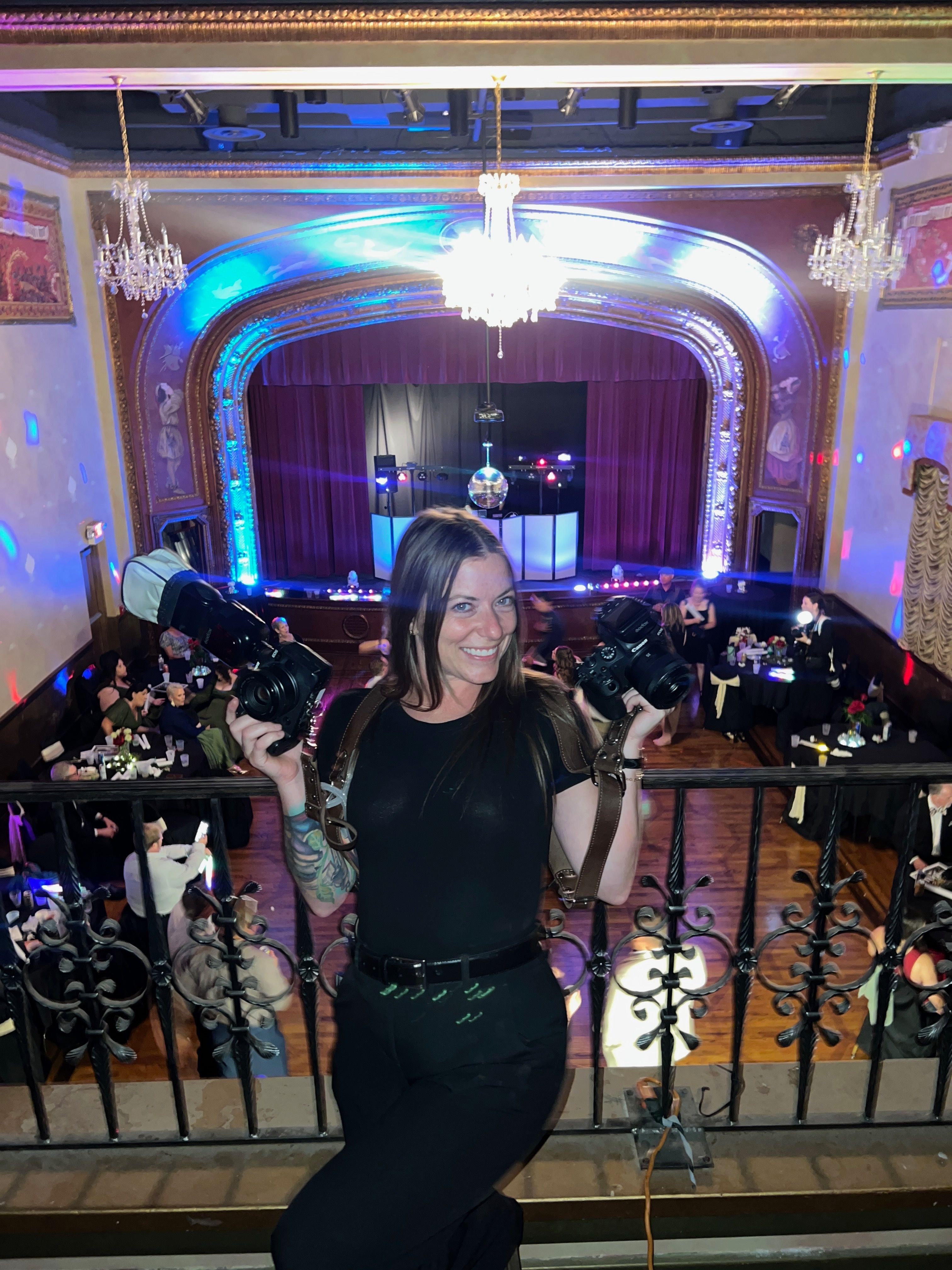 Woman with tattoos and long hair holding a drink, standing near a railing at a decorated event with blue lighting.