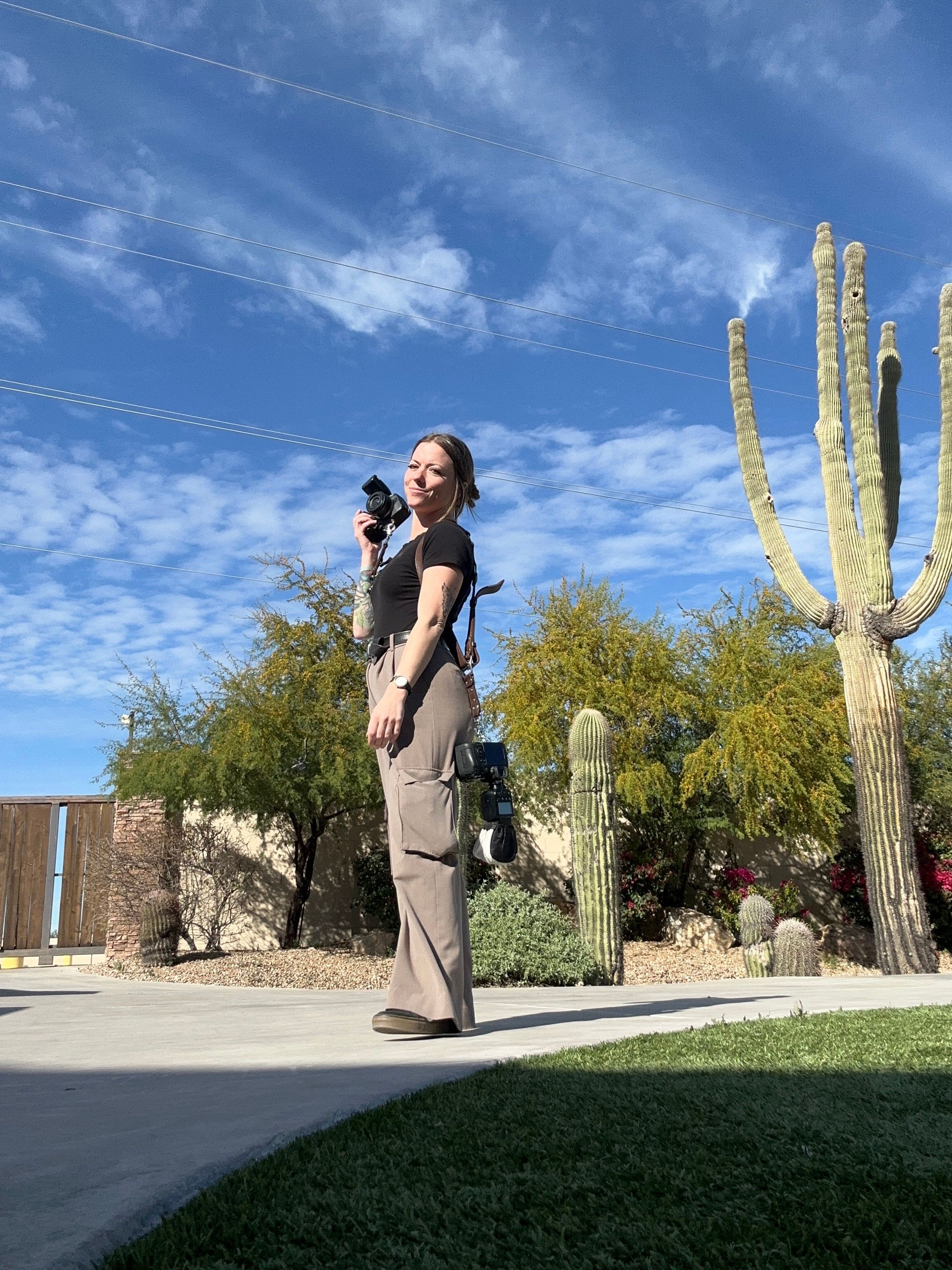 Woman standing outdoors near desert plants, holding a camera, with a large cactus and blue sky in background.