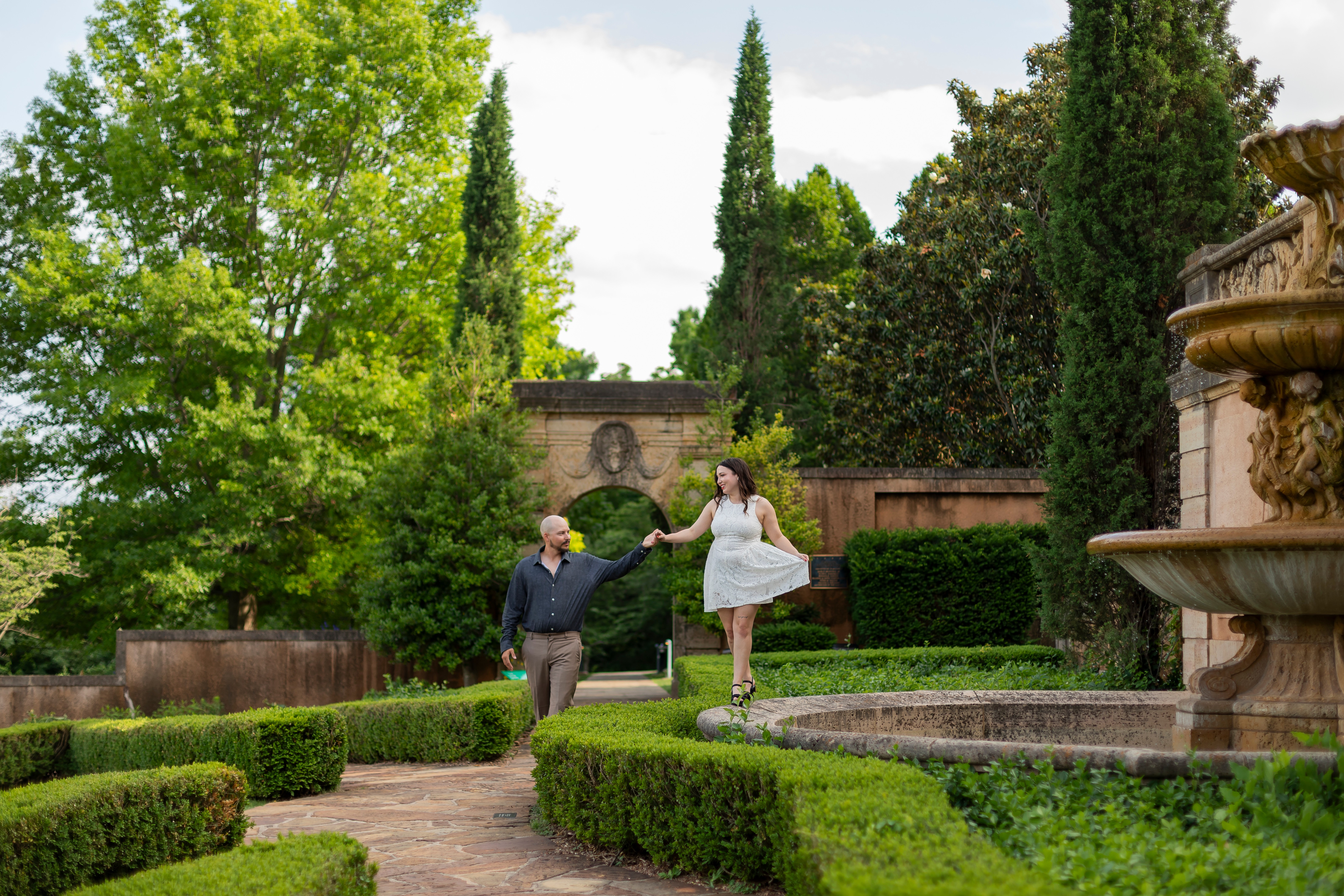 Two people, one in a white dress and the other in dark clothing, walk in a lush garden with trees and a fountain.