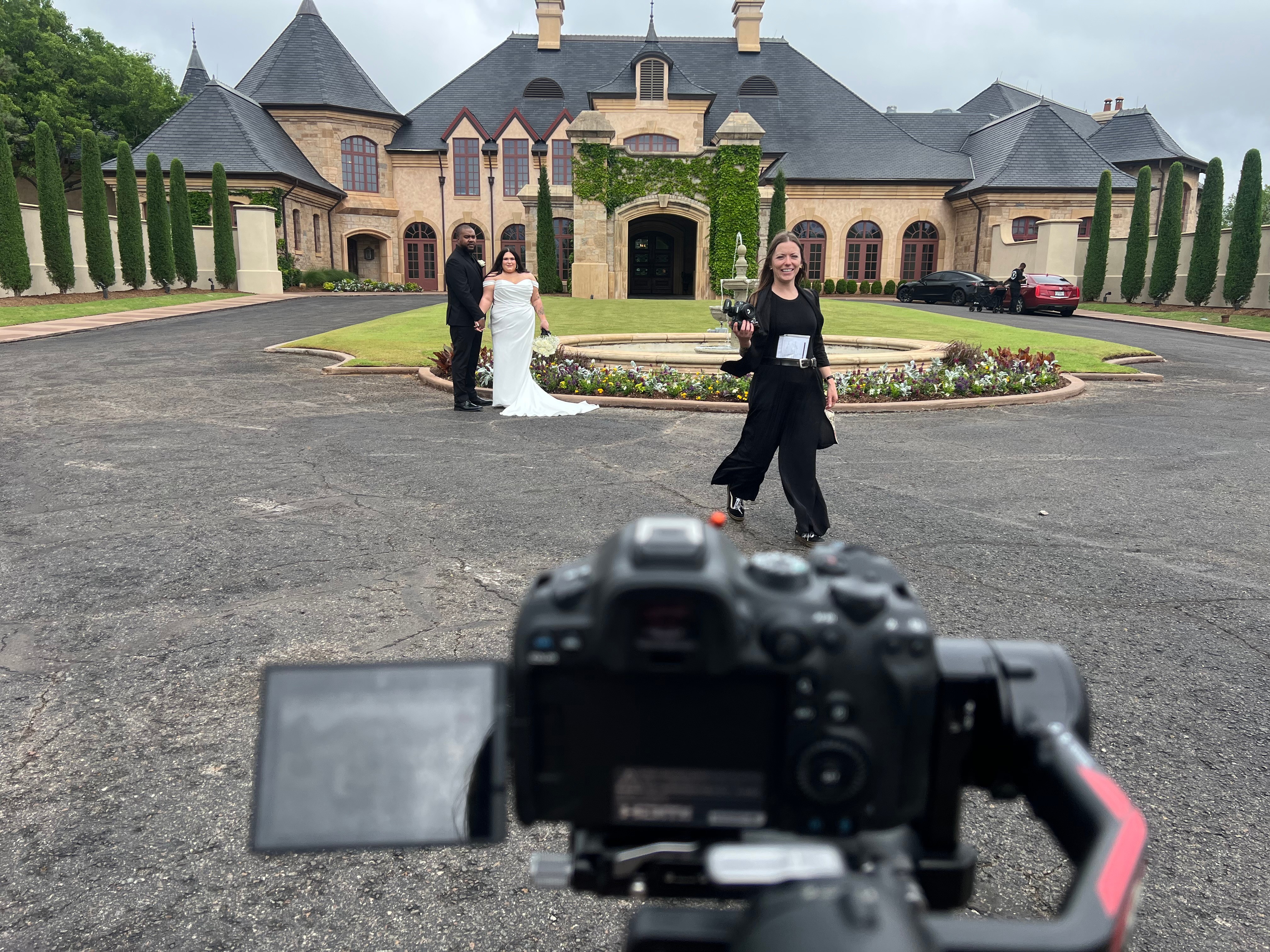Person standing in front of a large mansion with a group of people, camera in foreground, cloudy sky.