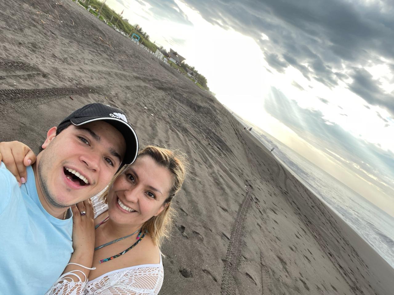 A smiling man and woman taking a selfie on a beach with cloudy sky and shoreline in background.