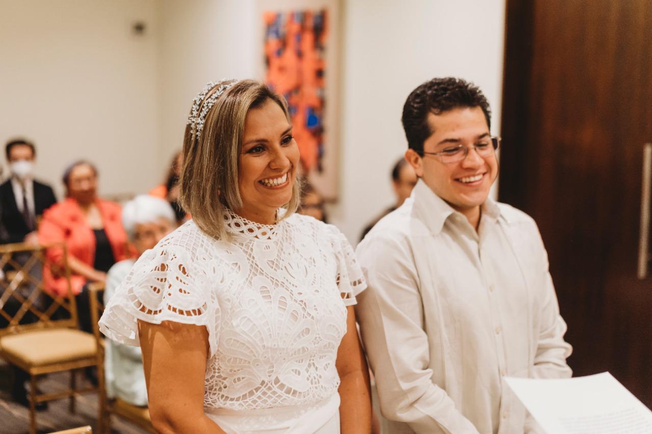 Two smiling people, a woman with a tiara and a man, stand in a room with seated guests in the background.