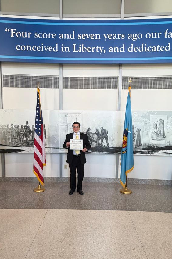 Person holding a certificate standing between American and Minnesota flags in front of a mural and a blue banner.