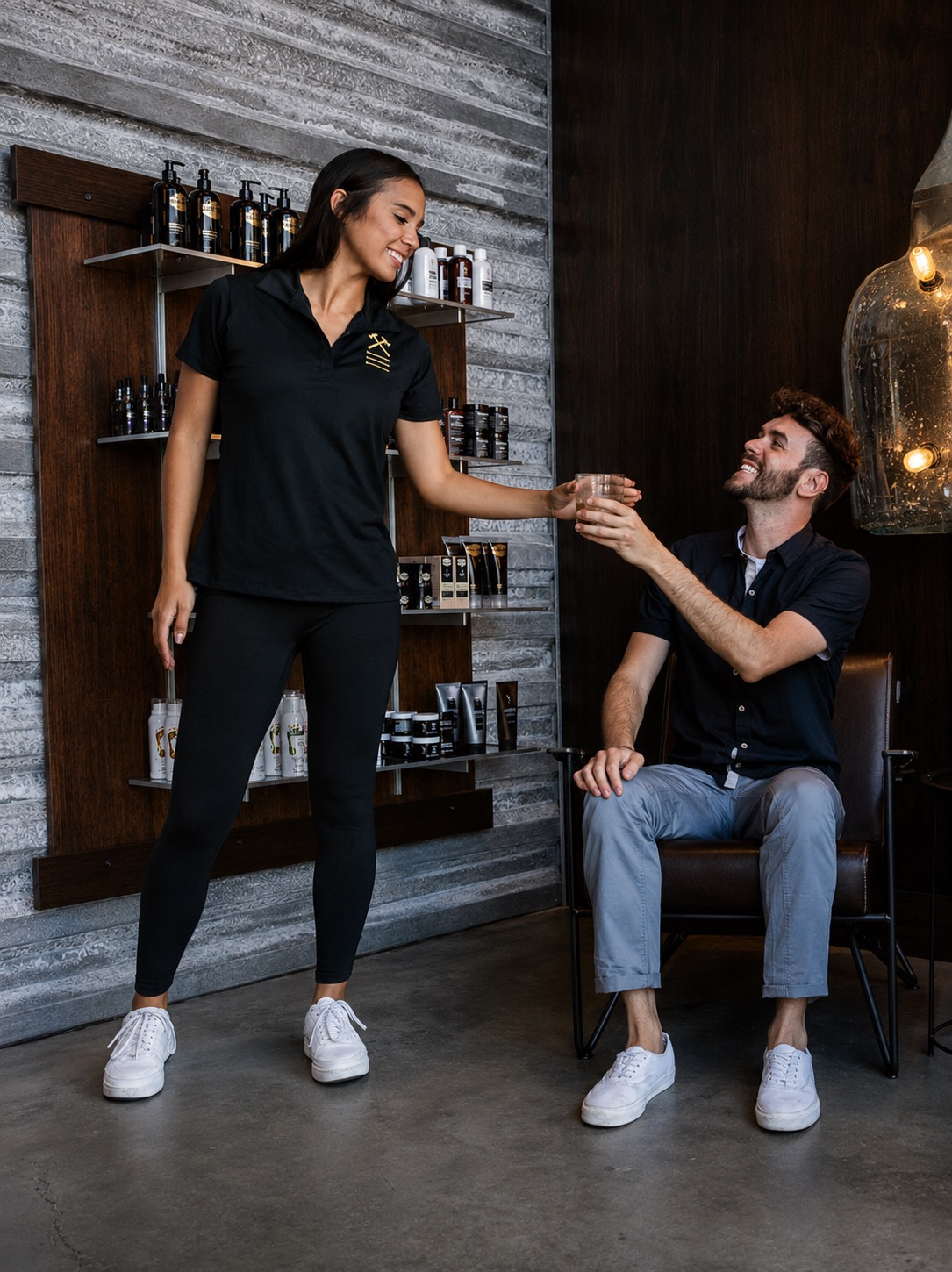 A woman in a black uniform hands a drink to a seated man in a cafe or bar setting, with shelves of bottles behind them.