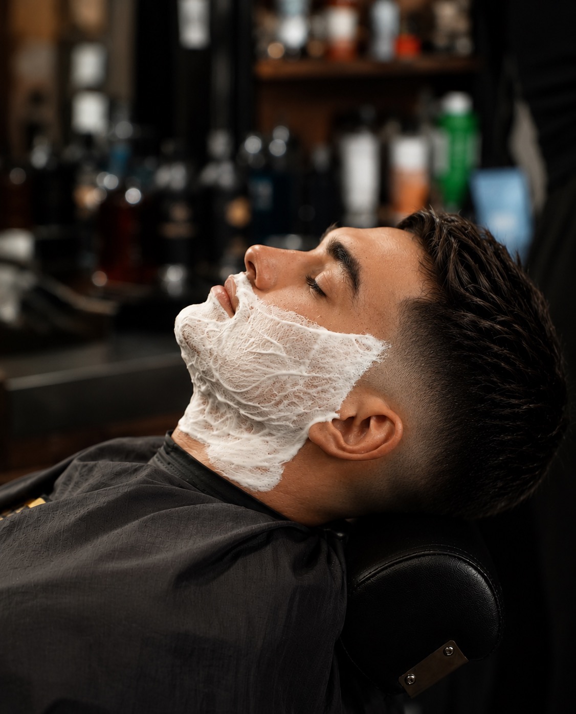 Person with a towel on face, lying back in a barber chair, in a barbershop setting.
