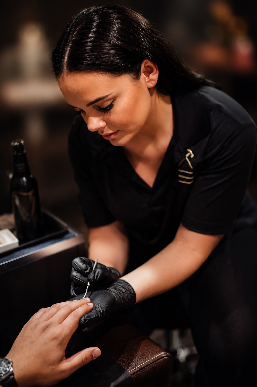 Woman with dark hair in a black uniform applies nail polish to a person's hand, wearing black gloves, in a salon setting.