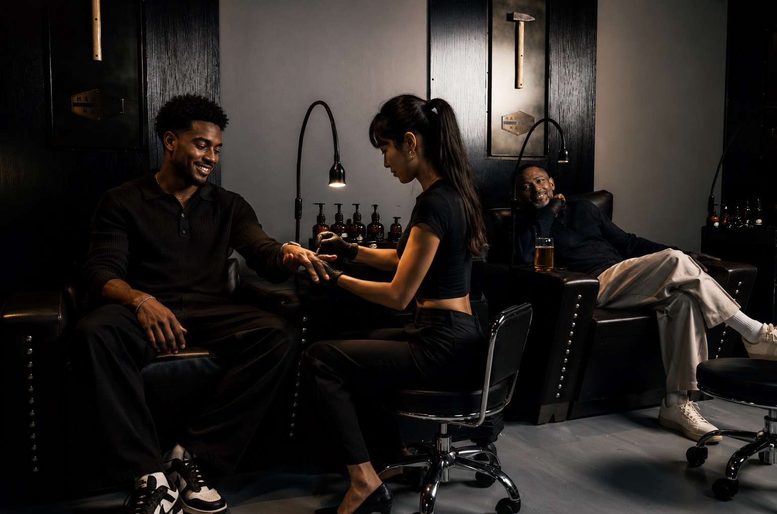 A woman is getting a manicure from a man in a dark room with black walls and lamps, while another person relaxes in the background.