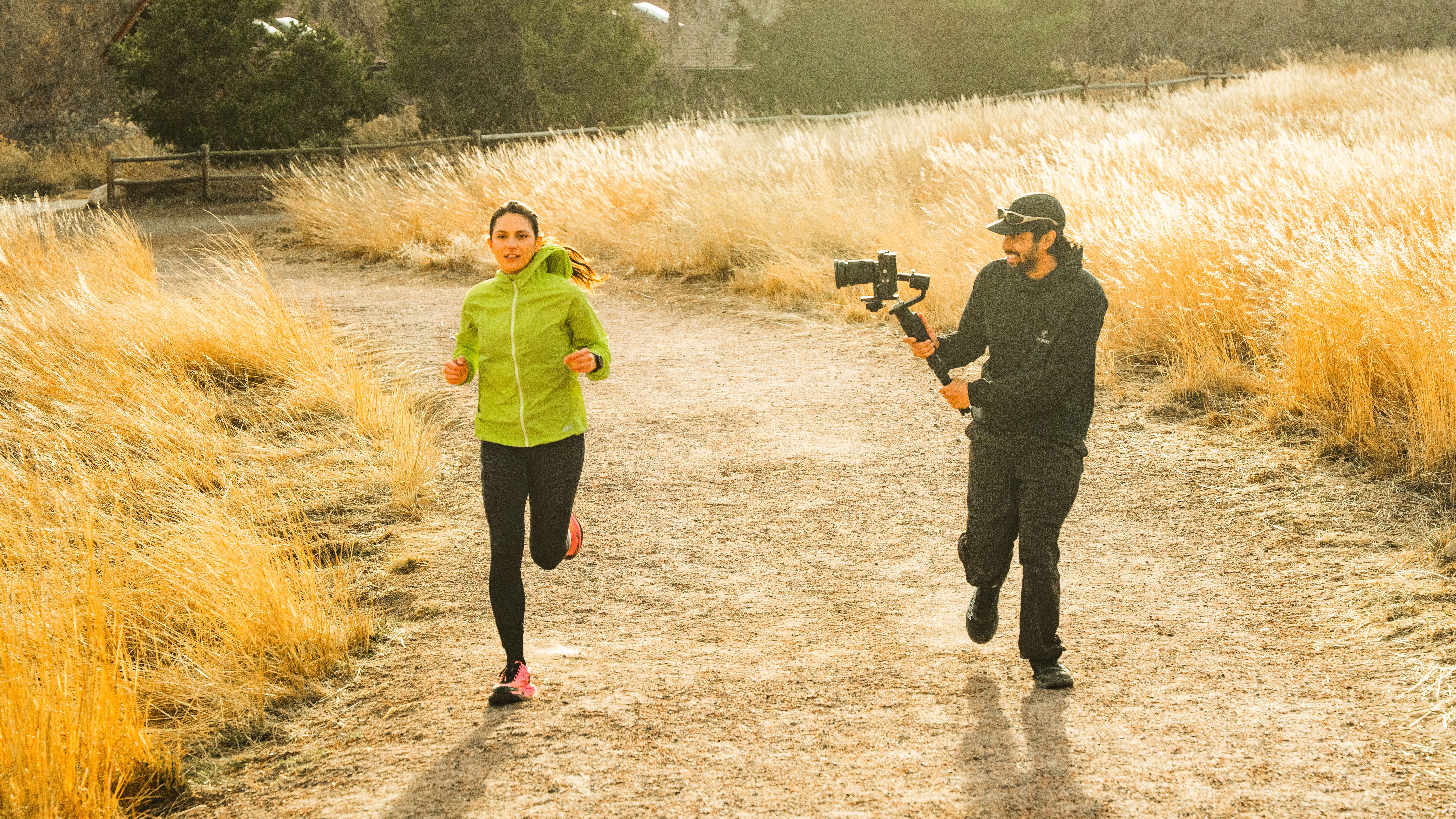 A woman runs on a dirt path with tall yellow grass on both sides, while a person films her with a camera.