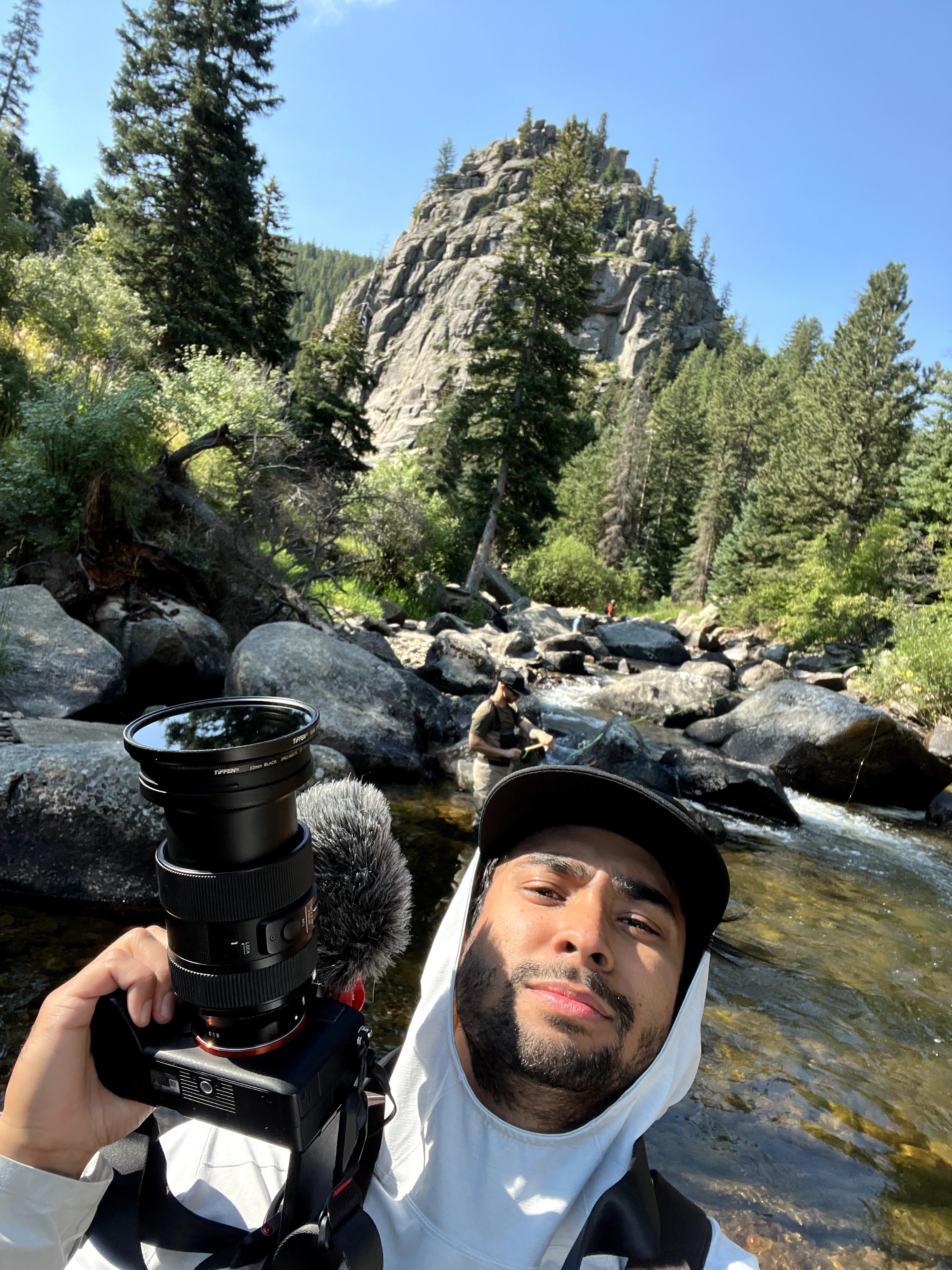 Man with a beard wearing a white hoodie and black cap, holding a camera, standing near a river with rocks and trees, mountain in background.