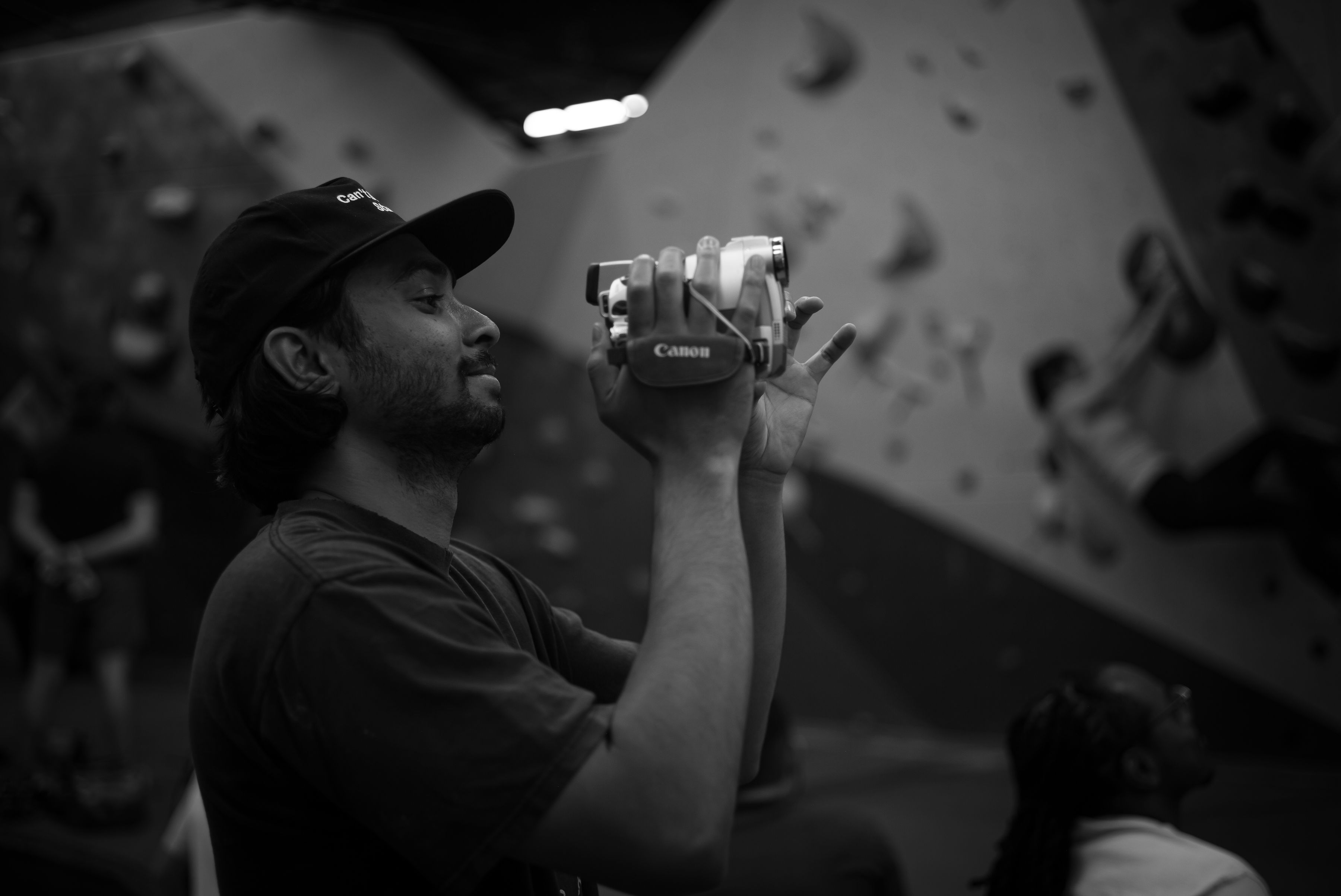 Person holding a camera, wearing a cap, with a climbing wall in the background, in black and white.