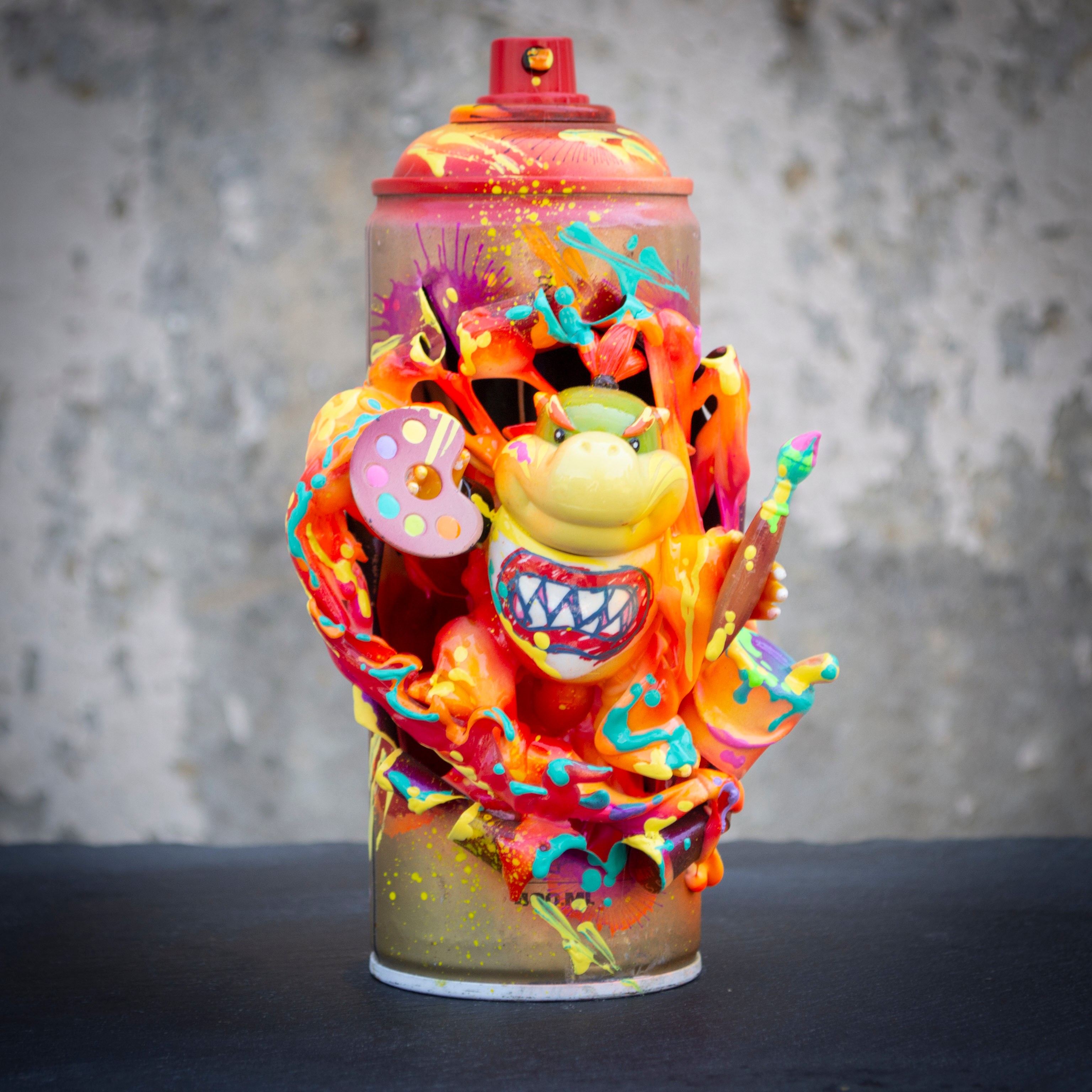 A spray paint can covered in colorful, abstract paint splashes and a cartoonish smiling face with a paint palette on its side.