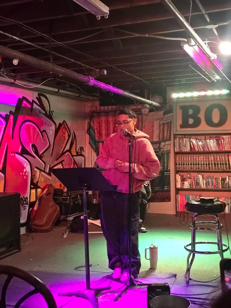 Person standing on stage with microphone, pink hoodie, in a room with graffiti and bookshelves, purple lighting, and a stool.