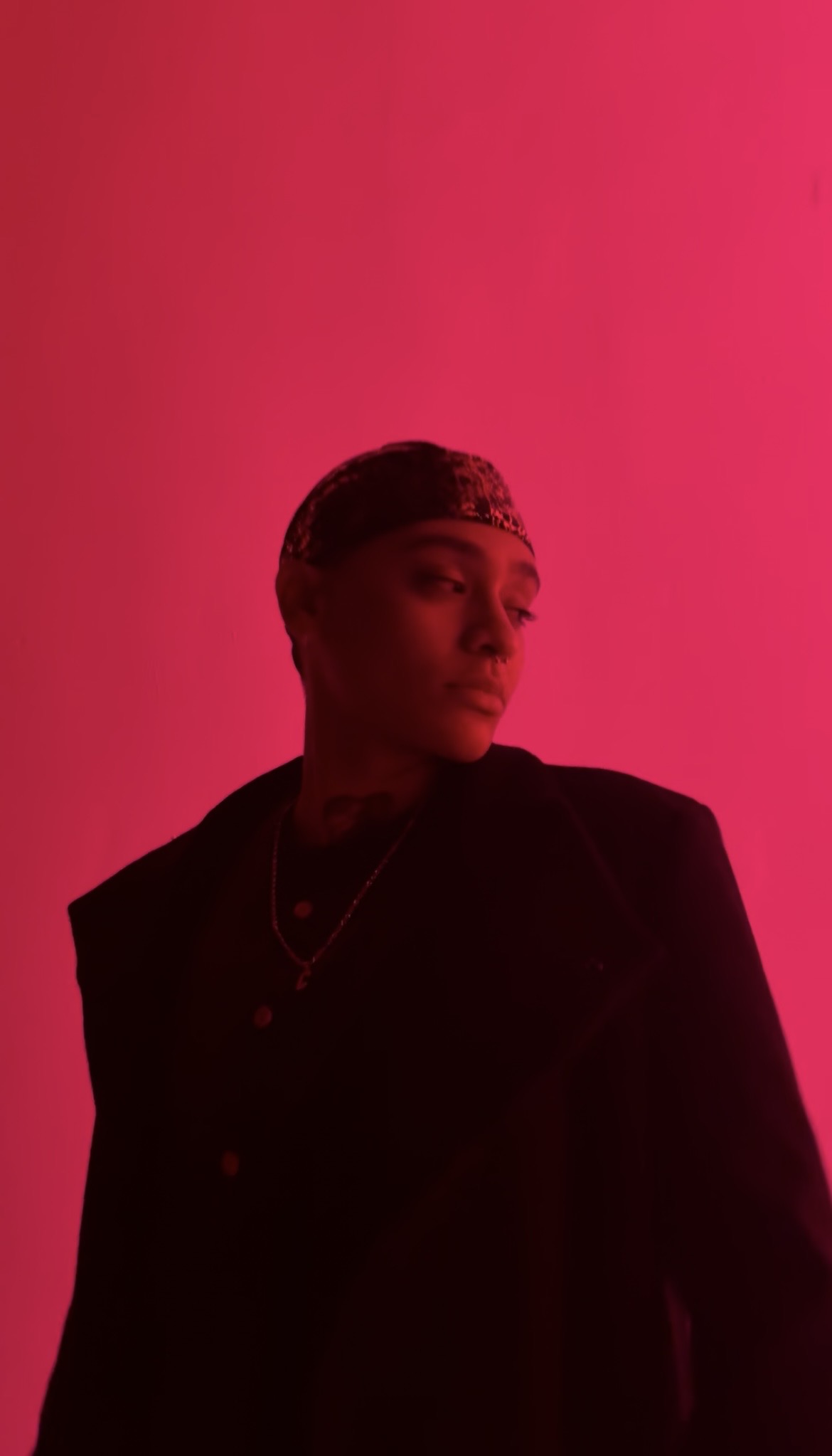 Person with short hair and necklace, wearing dark clothing, against a pink background, looking to the side.