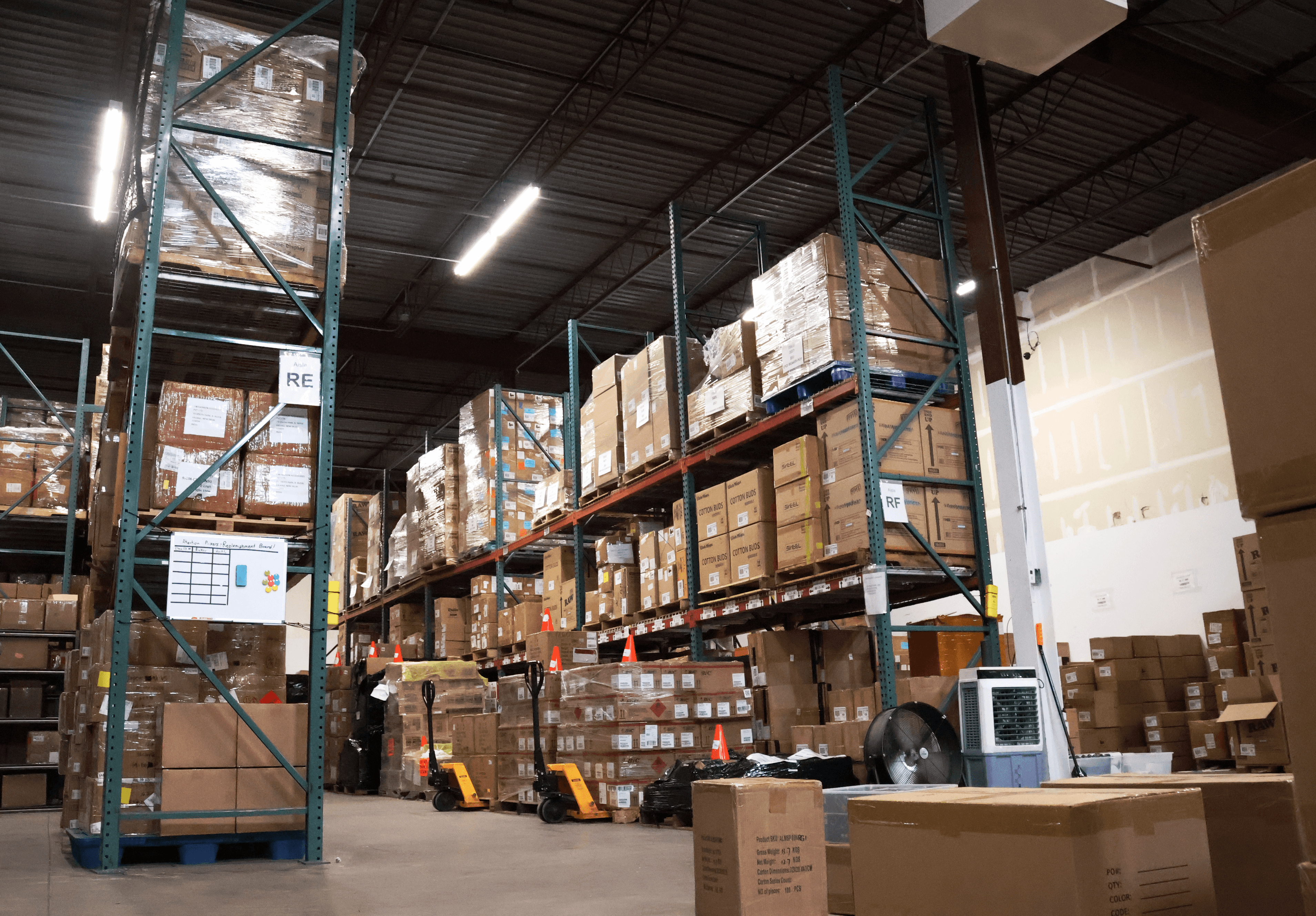 Warehouse with tall shelves filled with boxes and pallets, industrial lighting, and a concrete floor.