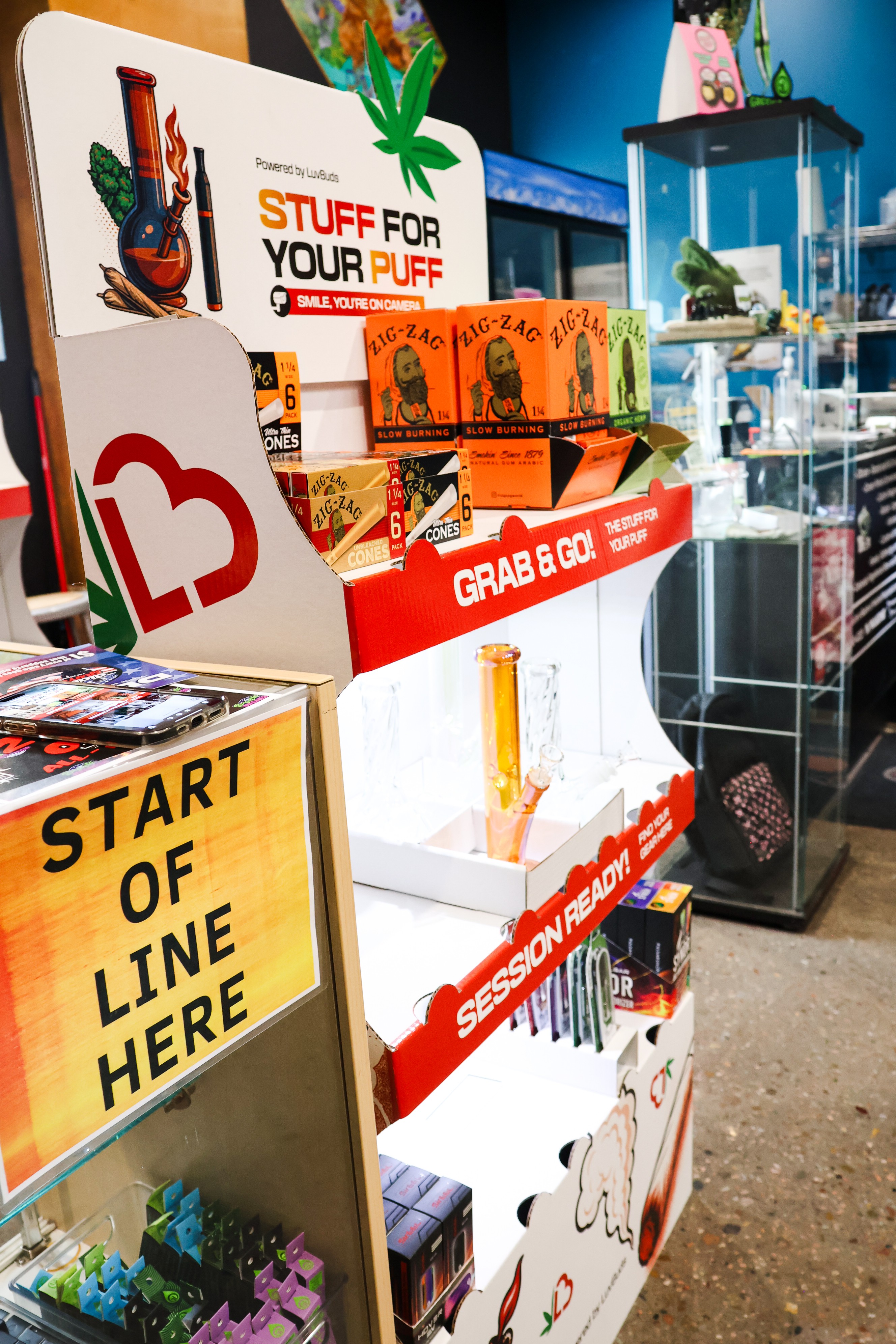 Display stand with cannabis-themed products and signs, including a yellow sign reading 'START OF LINE HERE,' in a retail store.