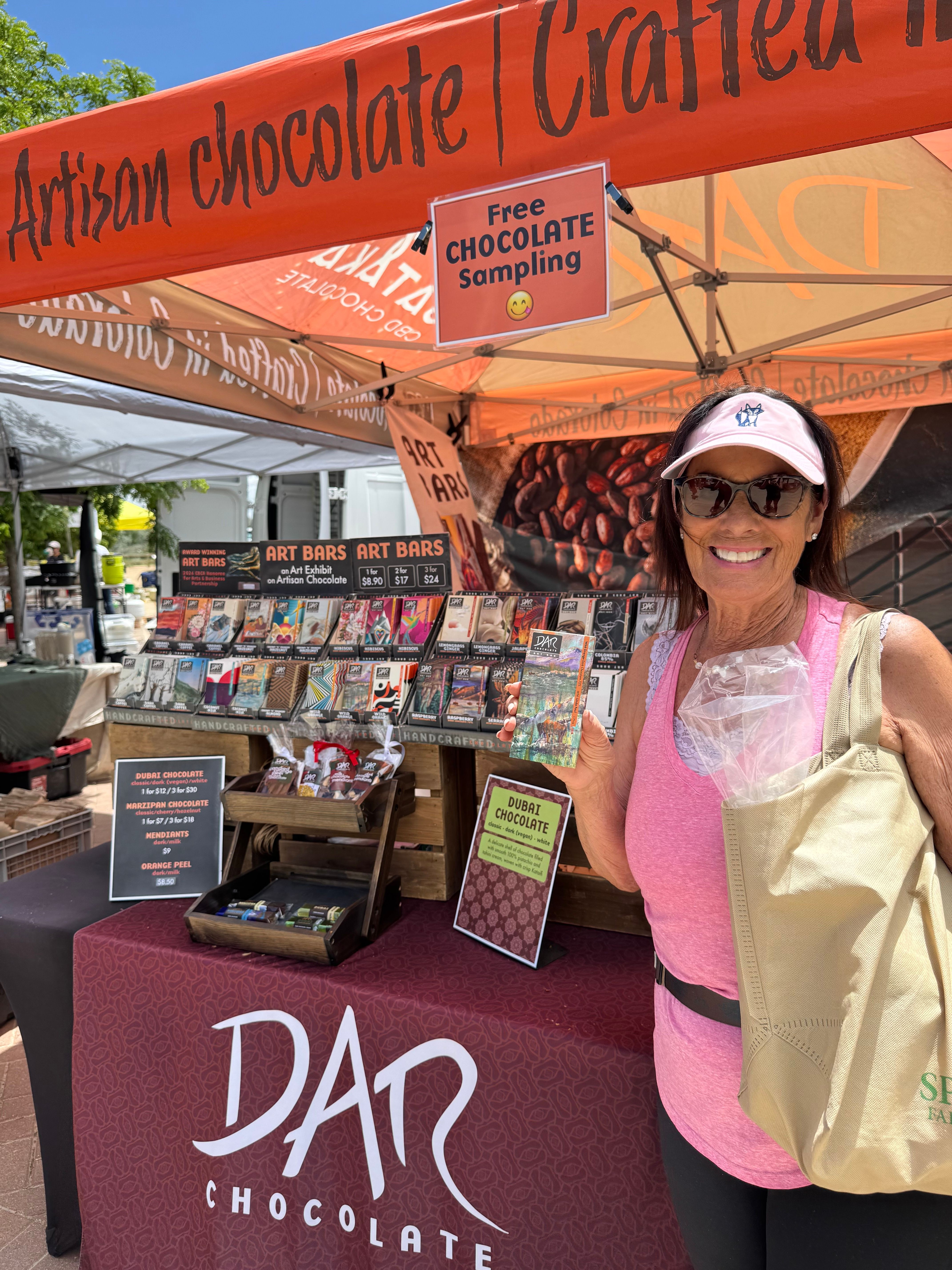 Woman with glasses and a pink shirt at a chocolate event, standing near a D'Art chocolate booth, with banners and products in background.