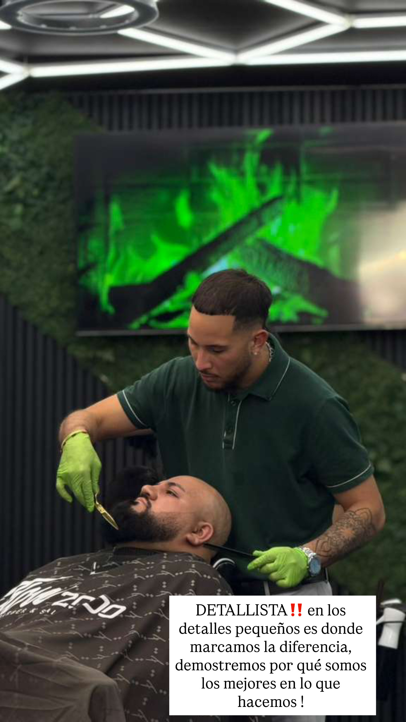 Barber applying shaving cream to a customer's face in a modern salon with a green-lit screen behind them.