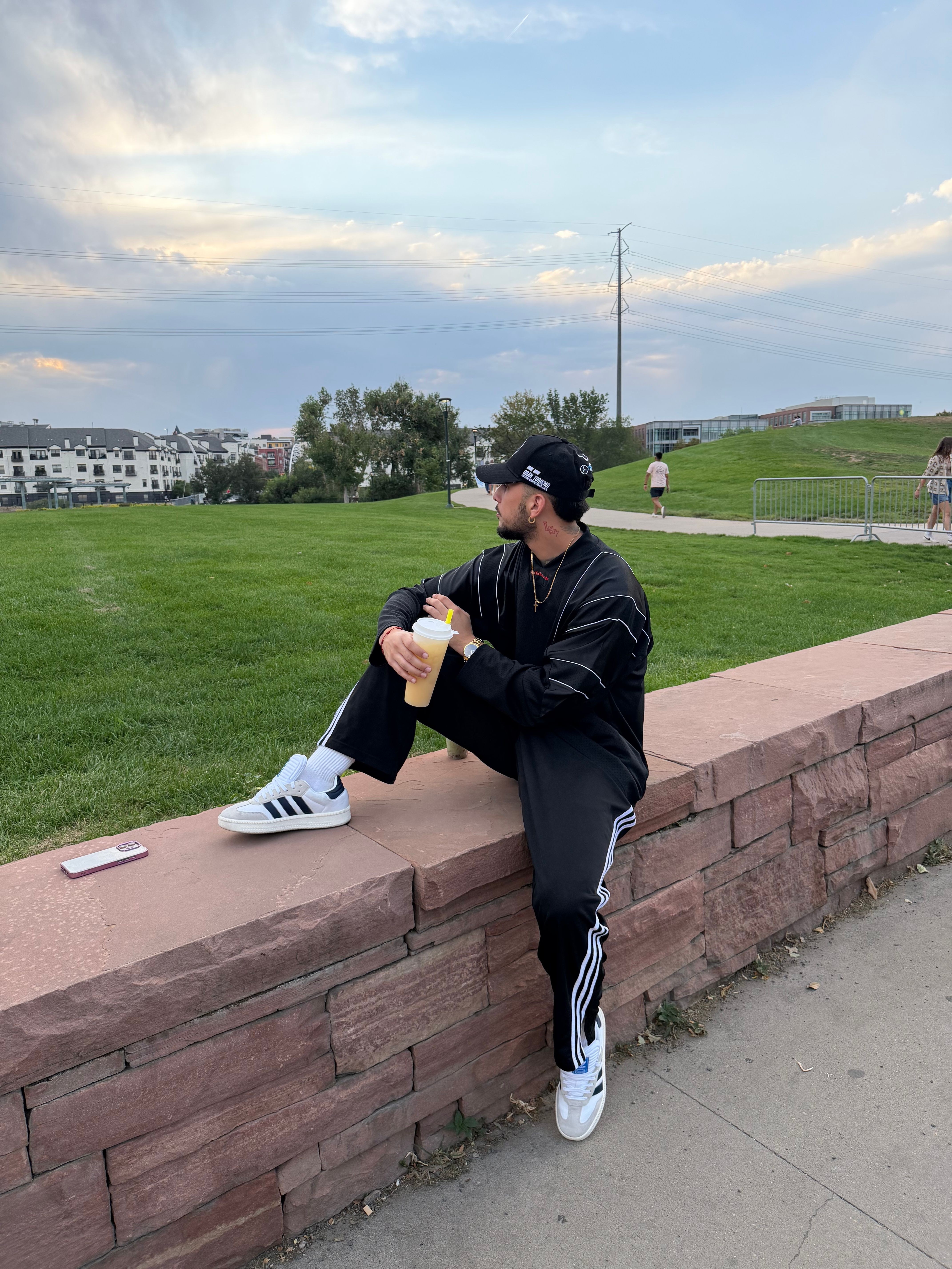 Person sitting on a brick ledge in a park, wearing a black jacket, black pants, and white sneakers, holding a yellow object.