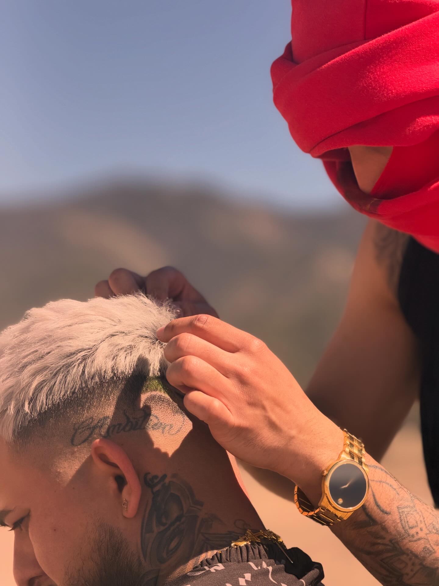 Person with short hair and tattoos adjusting hair of another person with a red headscarf outdoors.