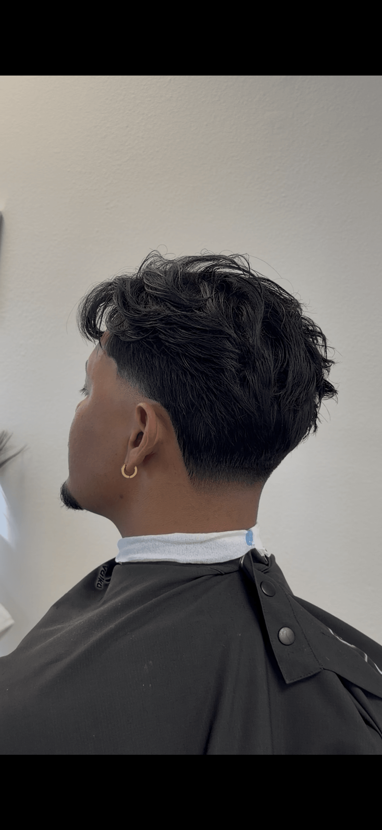 Side view of a person with short, wavy hair, wearing a black cape and a white collar, against a plain background.