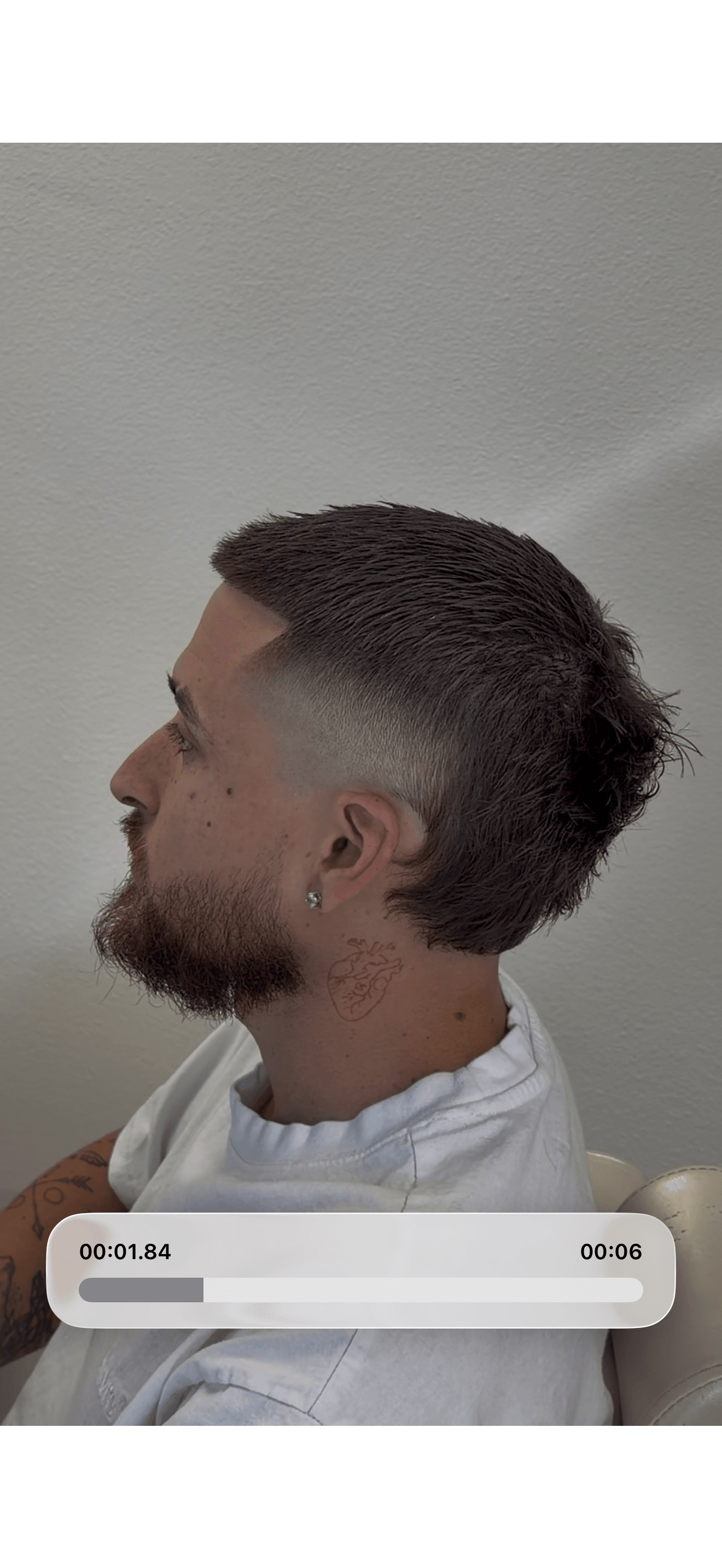 Profile of a man with short dark hair and beard, sitting against a plain wall, wearing a white shirt.