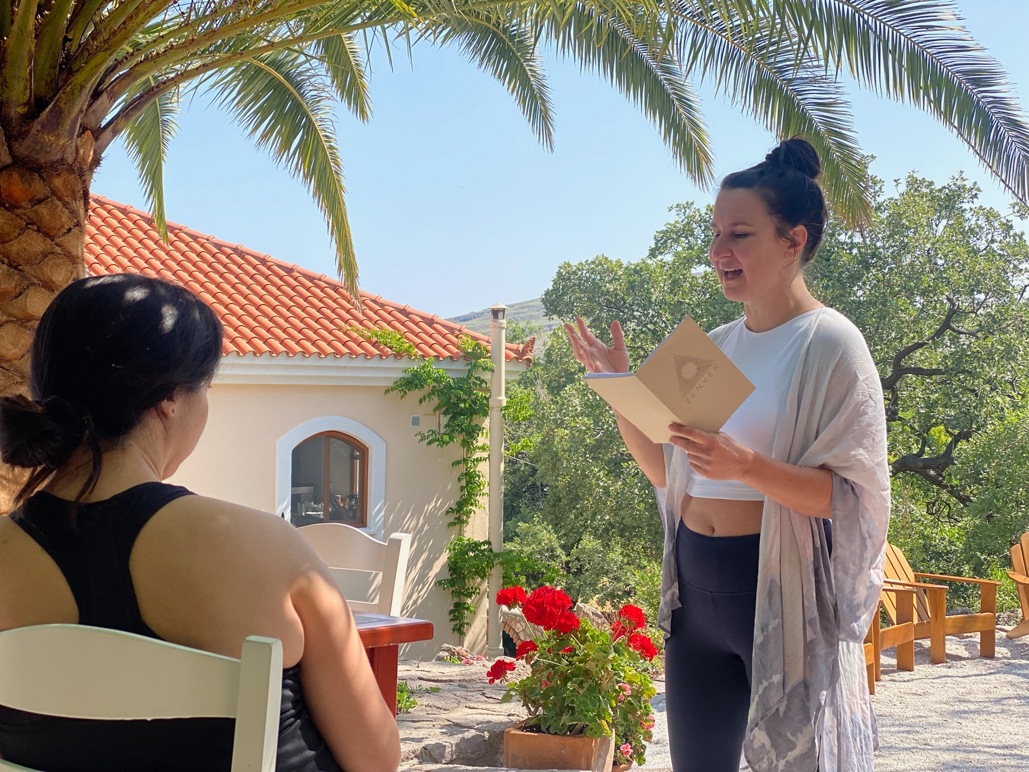 Two women talking outdoors under palm trees, one holding a book, with a building and flowers in background.