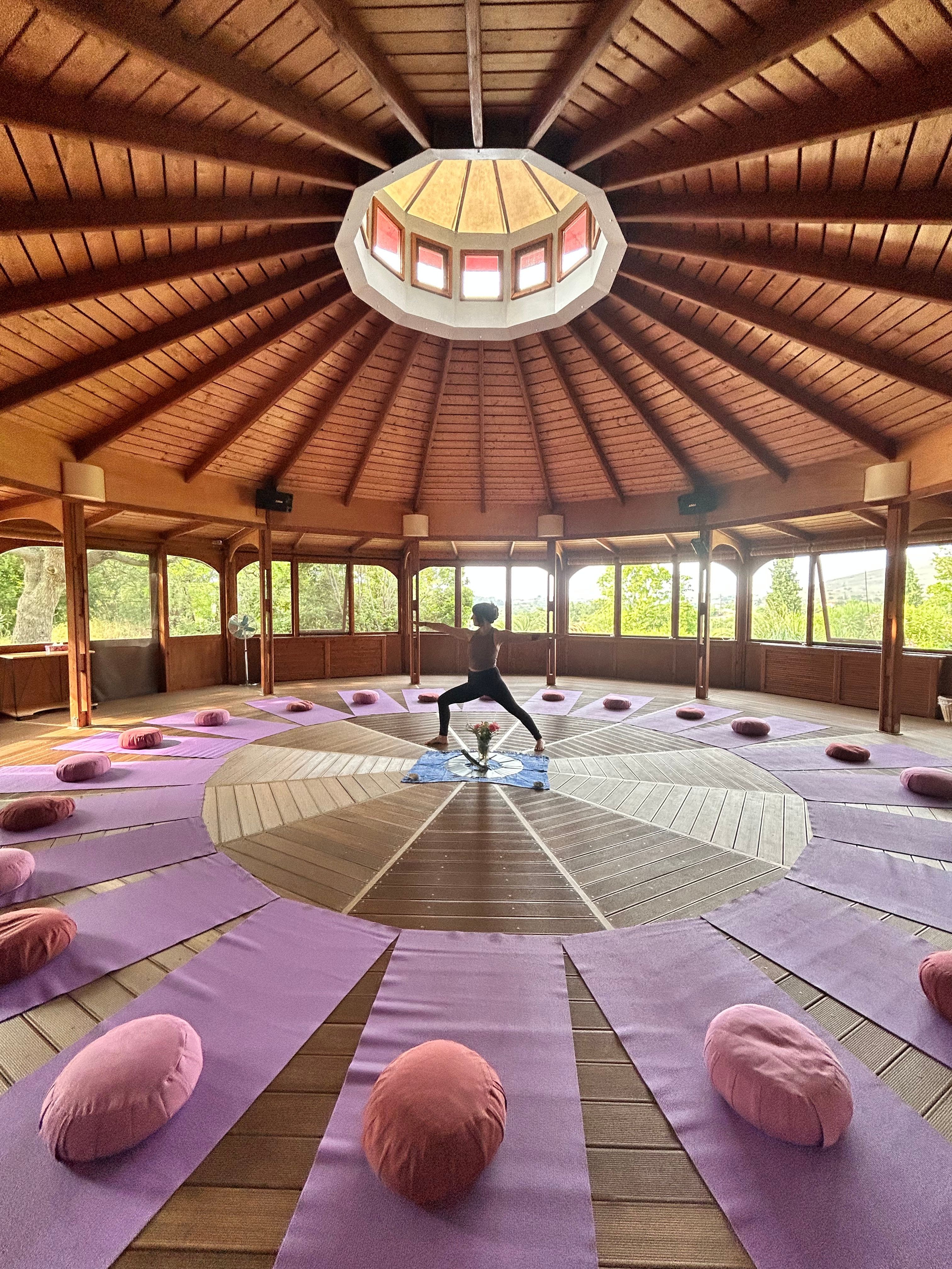 Person standing in a circular wooden room with purple cushions arranged around a central platform, large windows with trees outside.
