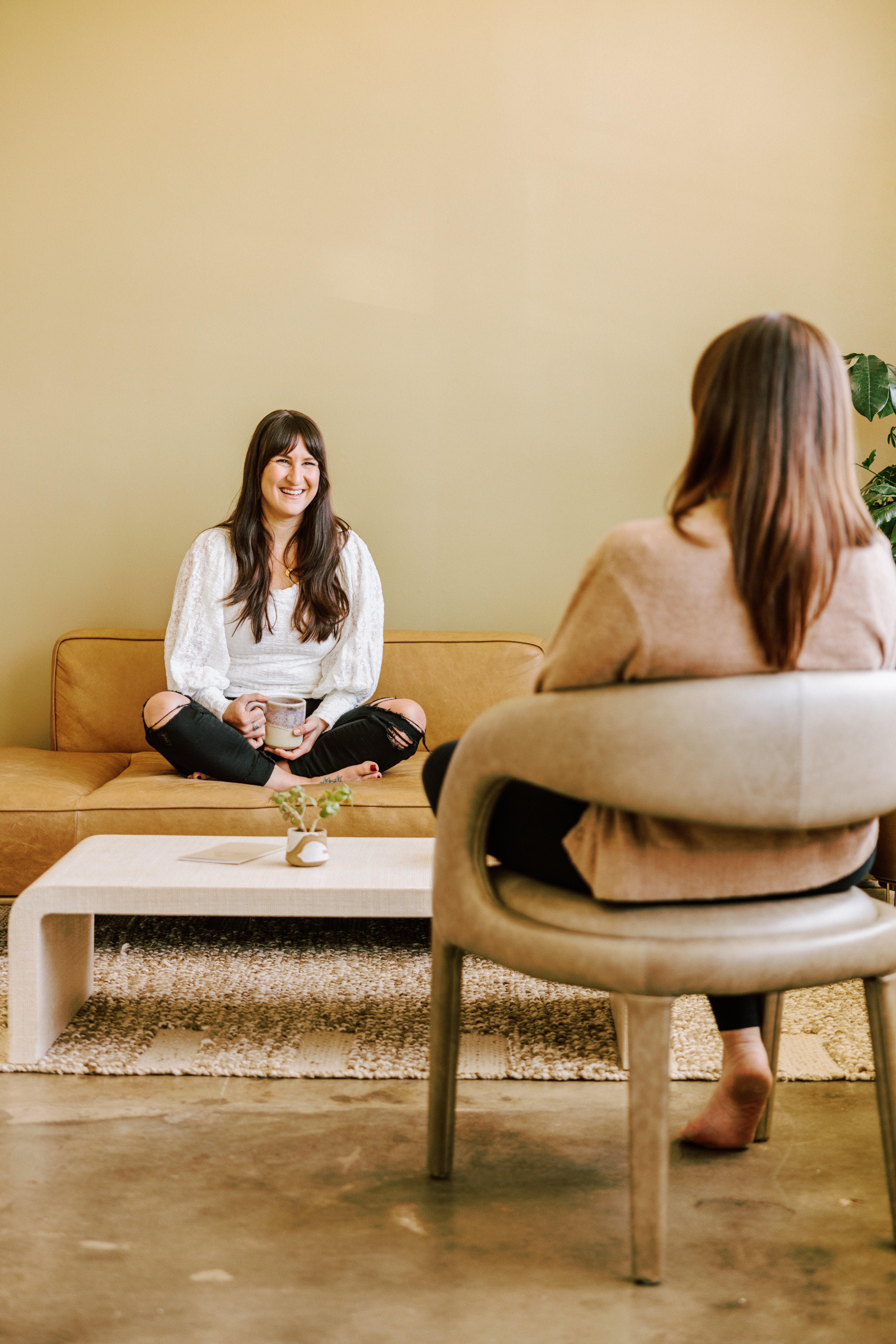 Two women sitting and talking in a room with a beige wall and modern furniture.