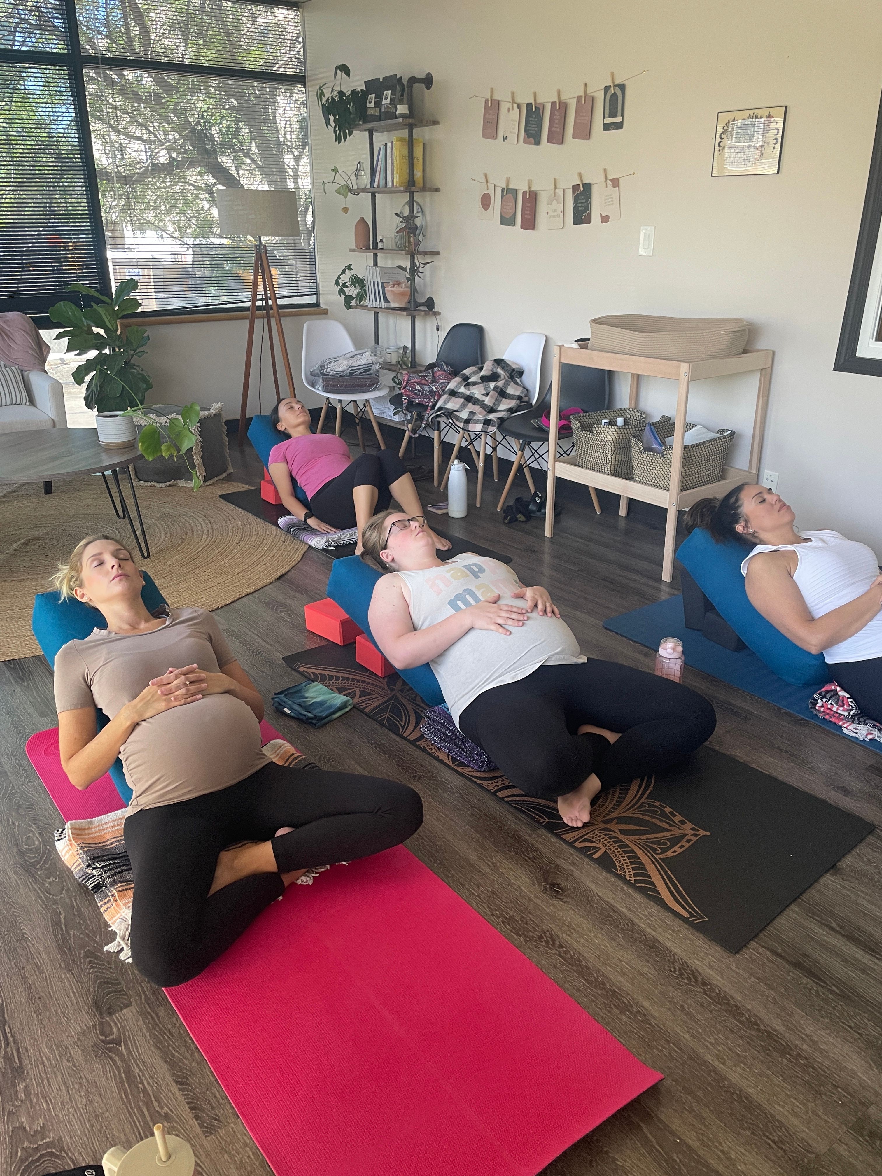 Two women practicing yoga on mats in a room with large windows, plants, and artwork, one sitting and one lying down.