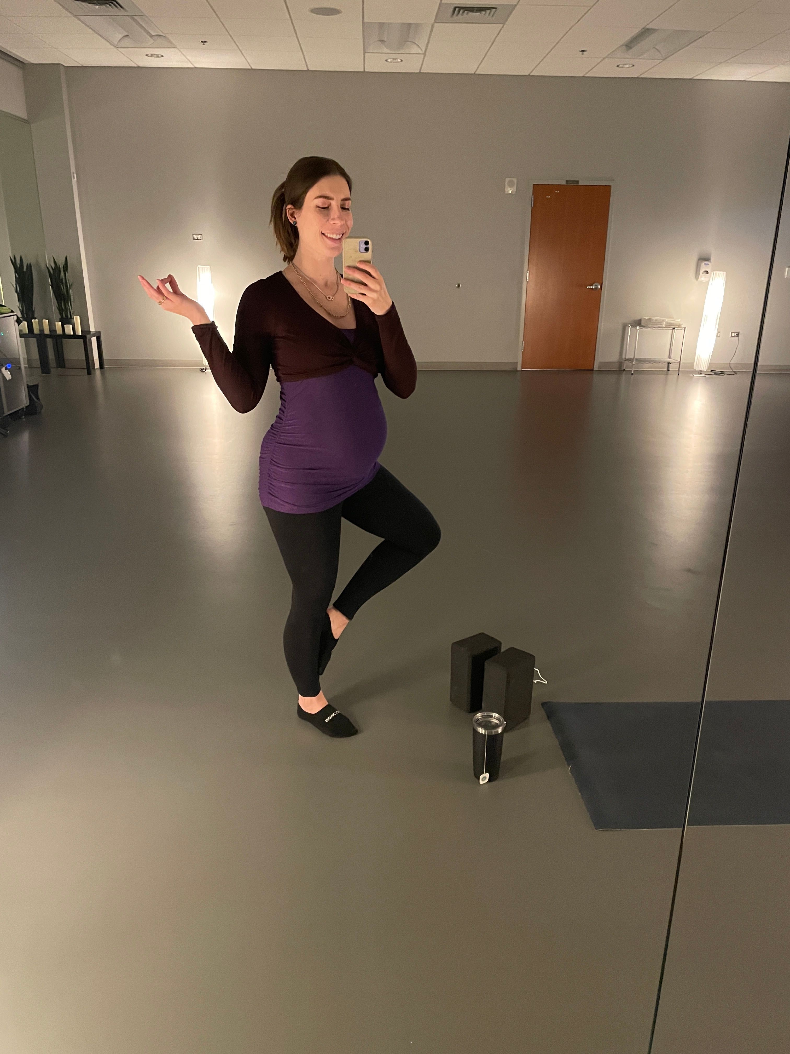 Pregnant woman in a purple top and black pants standing in a hallway, holding a phone to her ear.