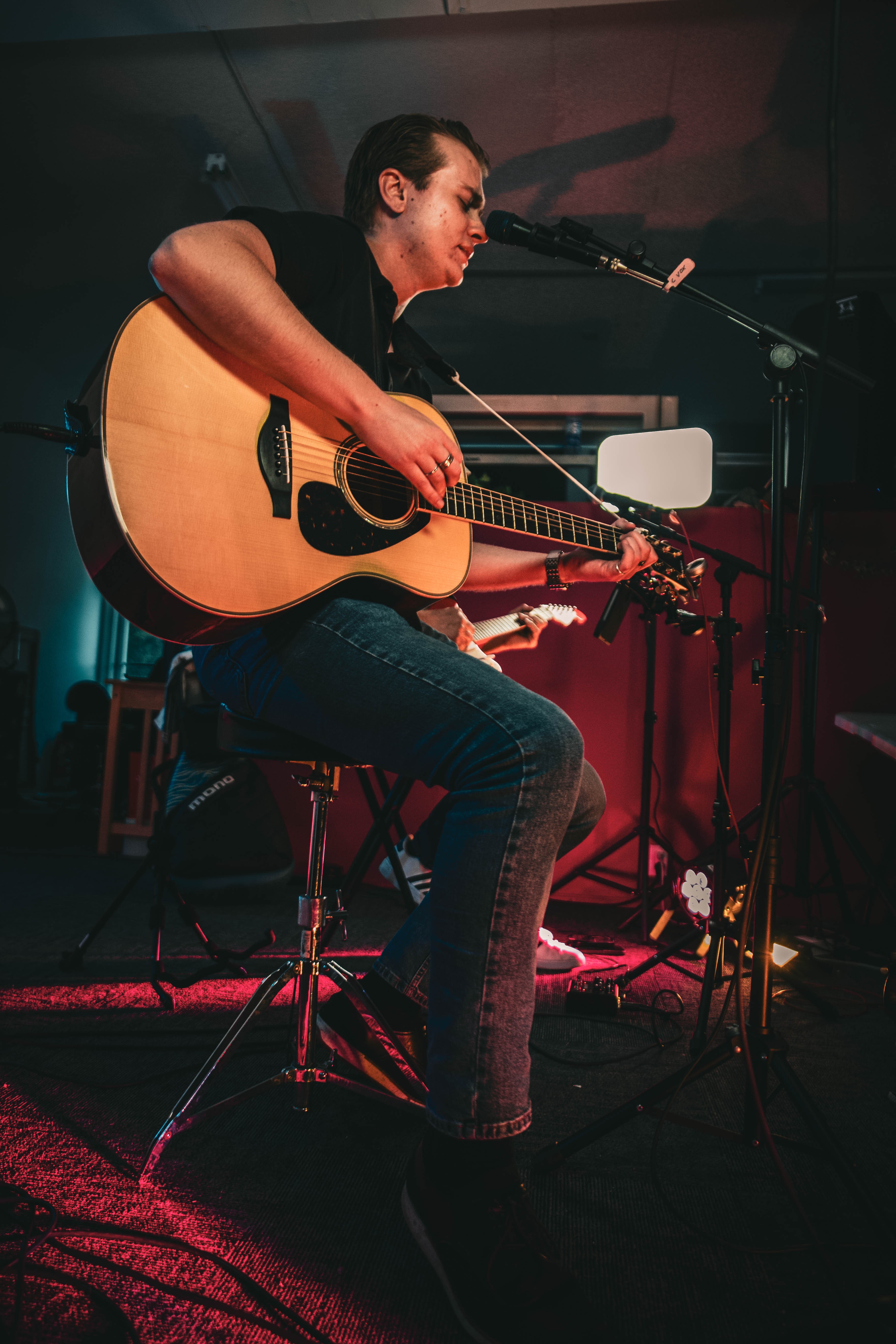 Person playing acoustic guitar and singing into a microphone on stage, with musical equipment around.